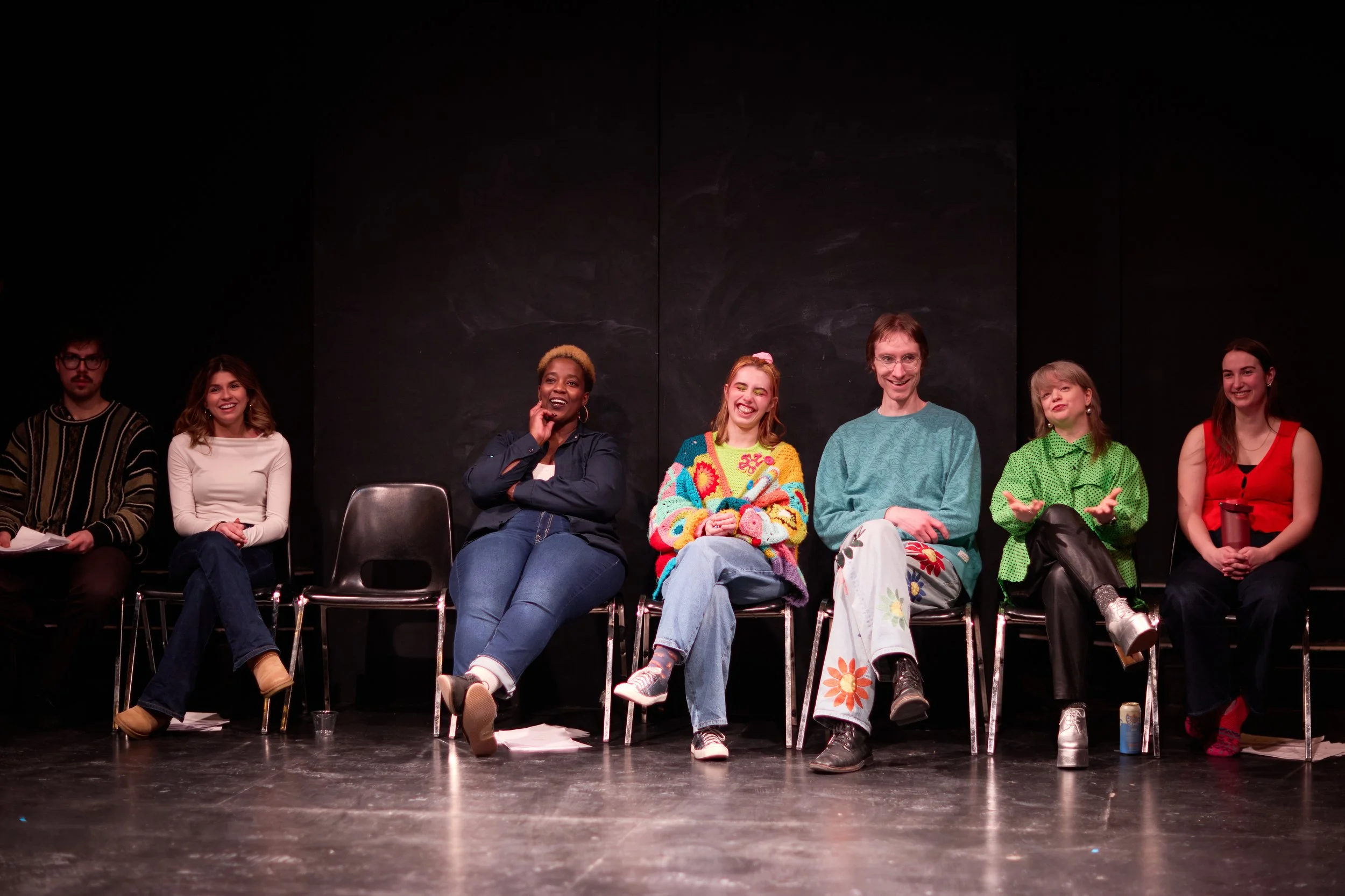 Sept personnes assises sur des chaises lors d'une pièce de théâtre ou d'un événement, riant et discutant, avec un mur noir en arrière-plan.