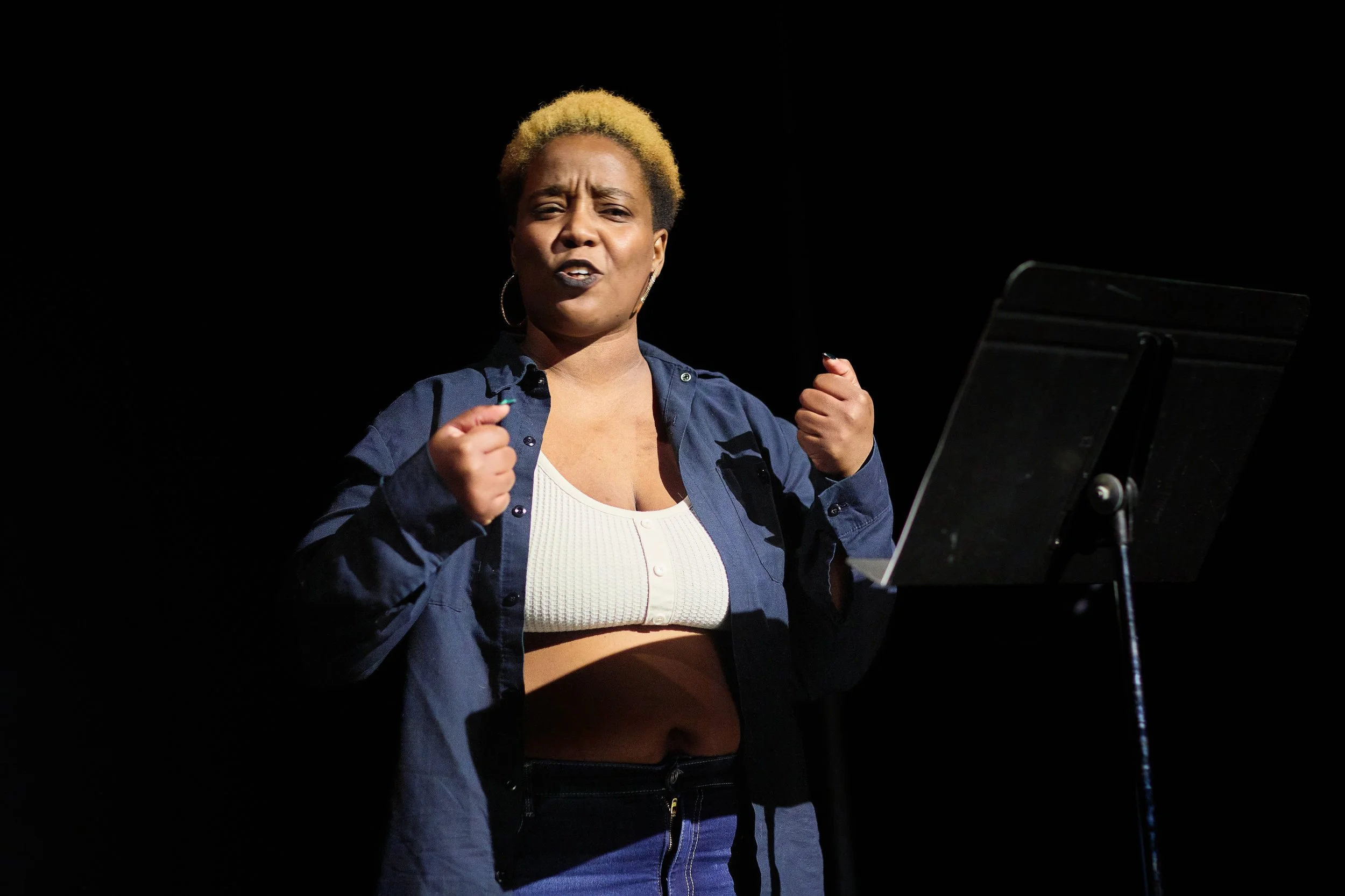 Une femme noire avec des cheveux blonds courts, portant un haut blanc, un jean et une chemise bleue, semble faire un discours expressif devant un pupitre, avec un fond noir.