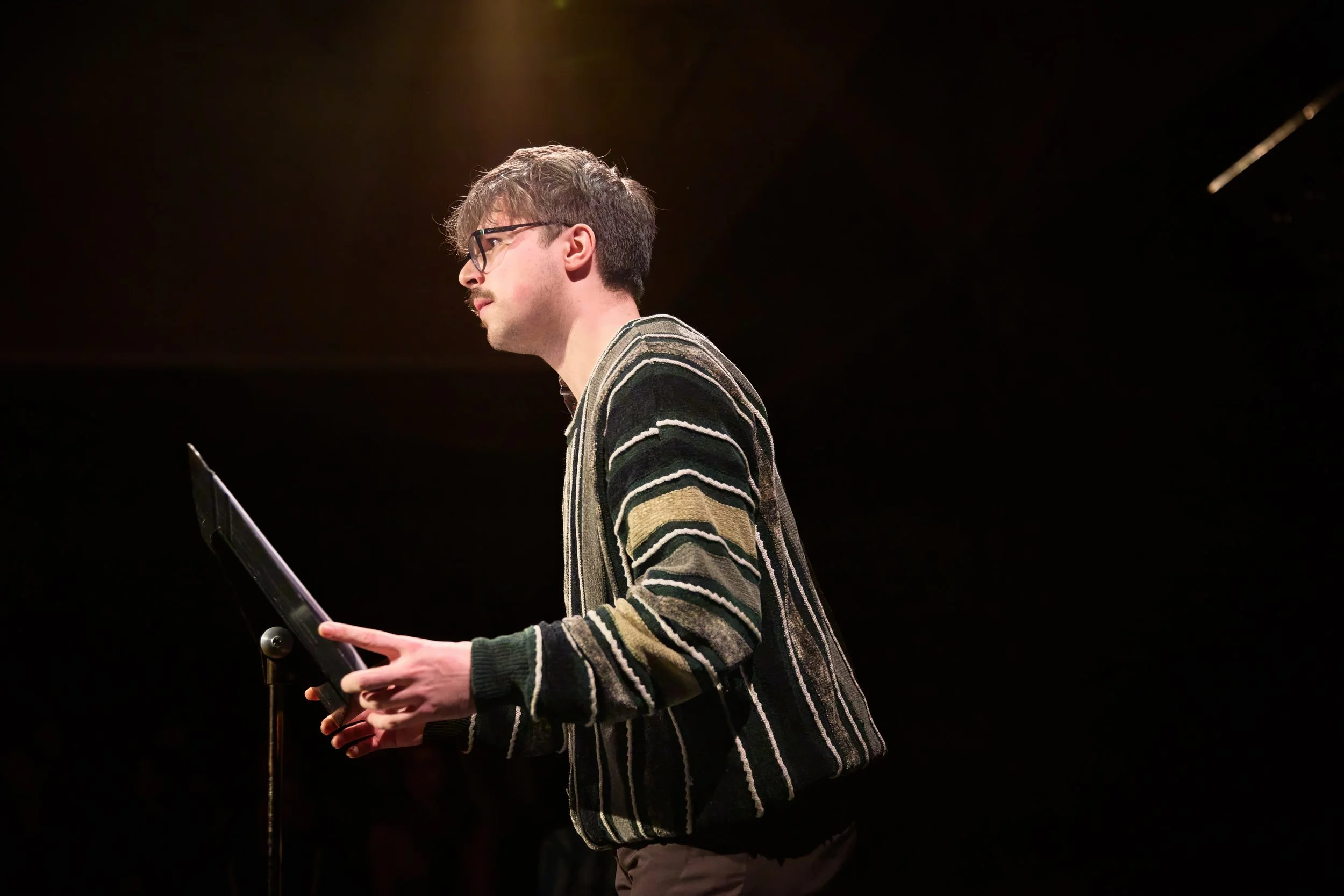 Jeune homme portant des lunettes, vêtu d'un pull à rayures, tenant une tablette, en train de parler ou de présenter sur scène, avec un fond noir.