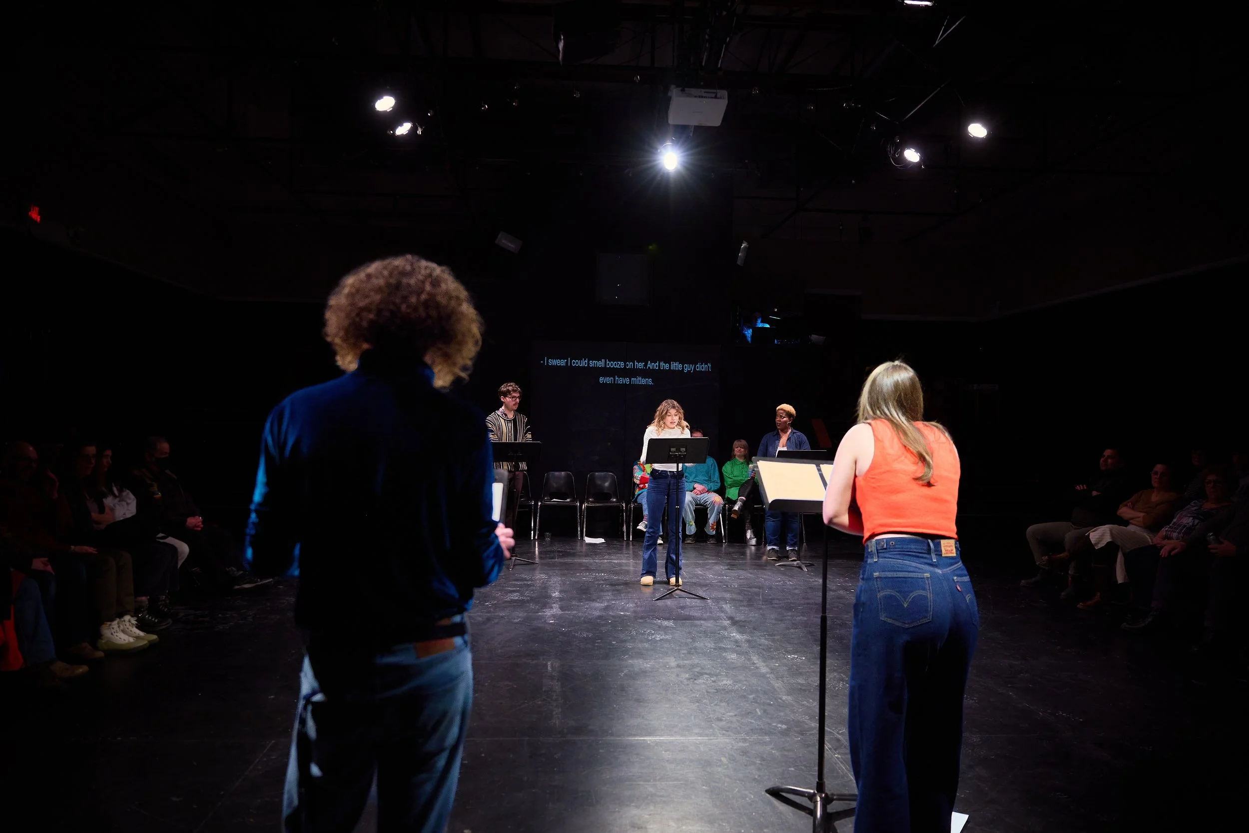 Groupe de personnes devant un micro sur scène lors d'une performance ou lecture, avec un public assis en demi-cercle dans une salle sombre. Projection de texte en arrière-plan.
