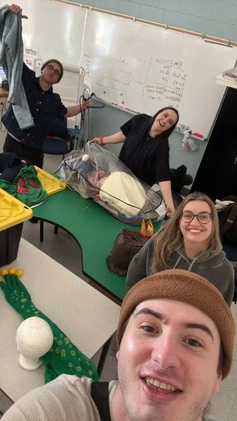 Trois jeunes femmes prennent un selfie dans une salle de classe devant un tableau blanc, avec un sac de matériel de bébé sur la table. Un ballon blanc et un haut en forme de crocodile vert à pois sont visibles au premier plan.