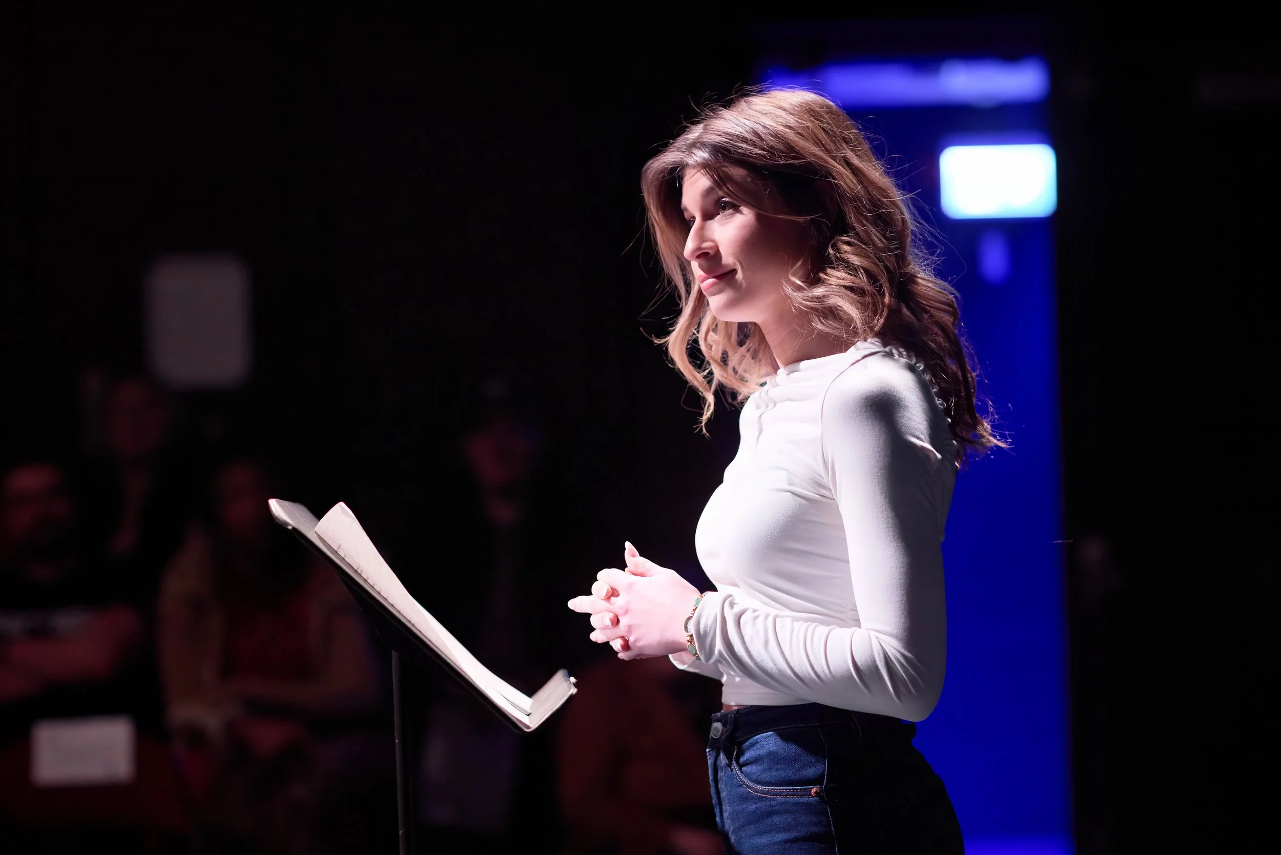 Une femme parle en public devant un pupitre, le public en arrière-plan est flou.