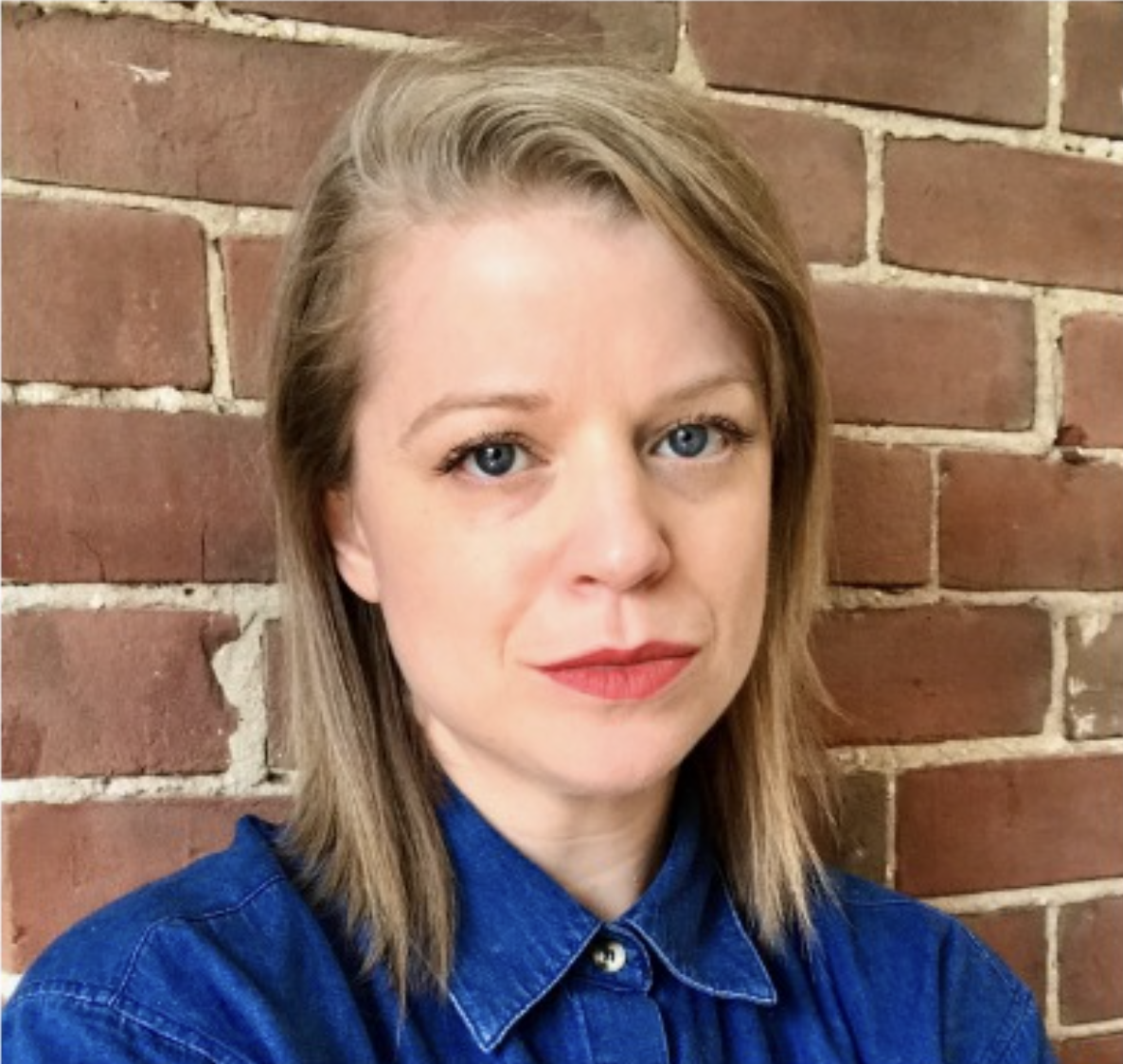 Portrait d'une femme avec des cheveux blonds et des yeux bleus, portant une chemise en denim bleu, devant un mur en briques