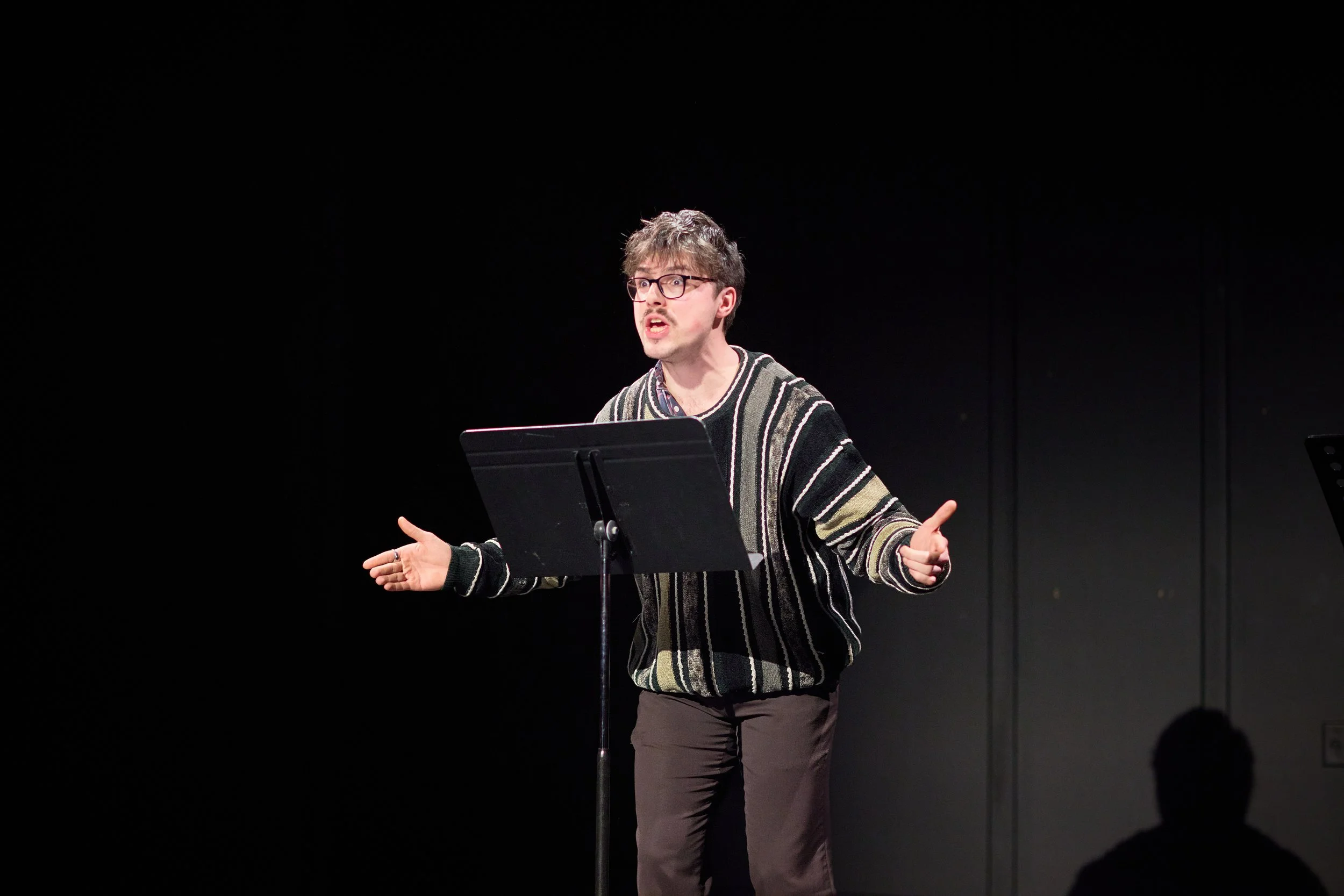 Un homme avec des lunettes, parlant et gesticulant lors d'une performance ou d'une lecture sur scène, avec un fond noir.
