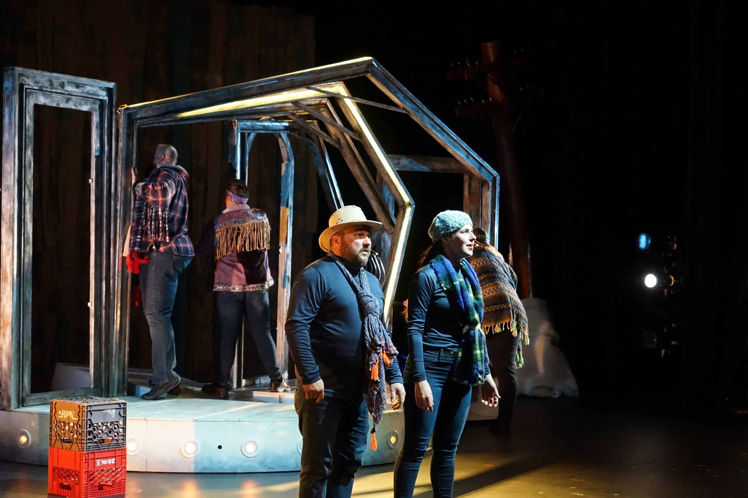 Scène de théâtre avec plusieurs acteurs en costume, certains portant des foulards ou des chapeaux, sur un décor de bois avec une structure géométrique lumineuse, en intérieur.