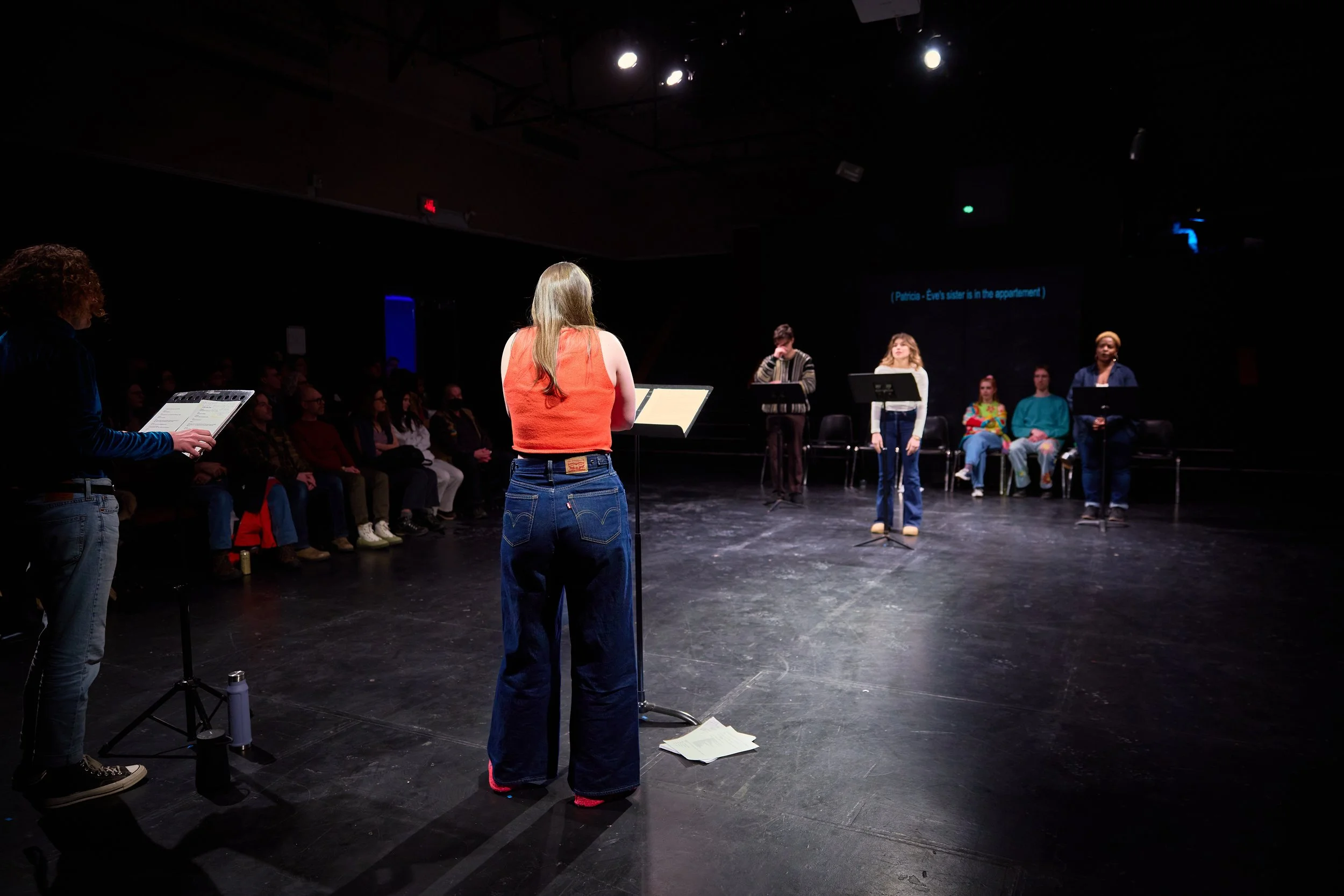 Scène de théâtre ou de répétition avec plusieurs personnes sur scène et un public assis, lumière focalisée sur les acteurs