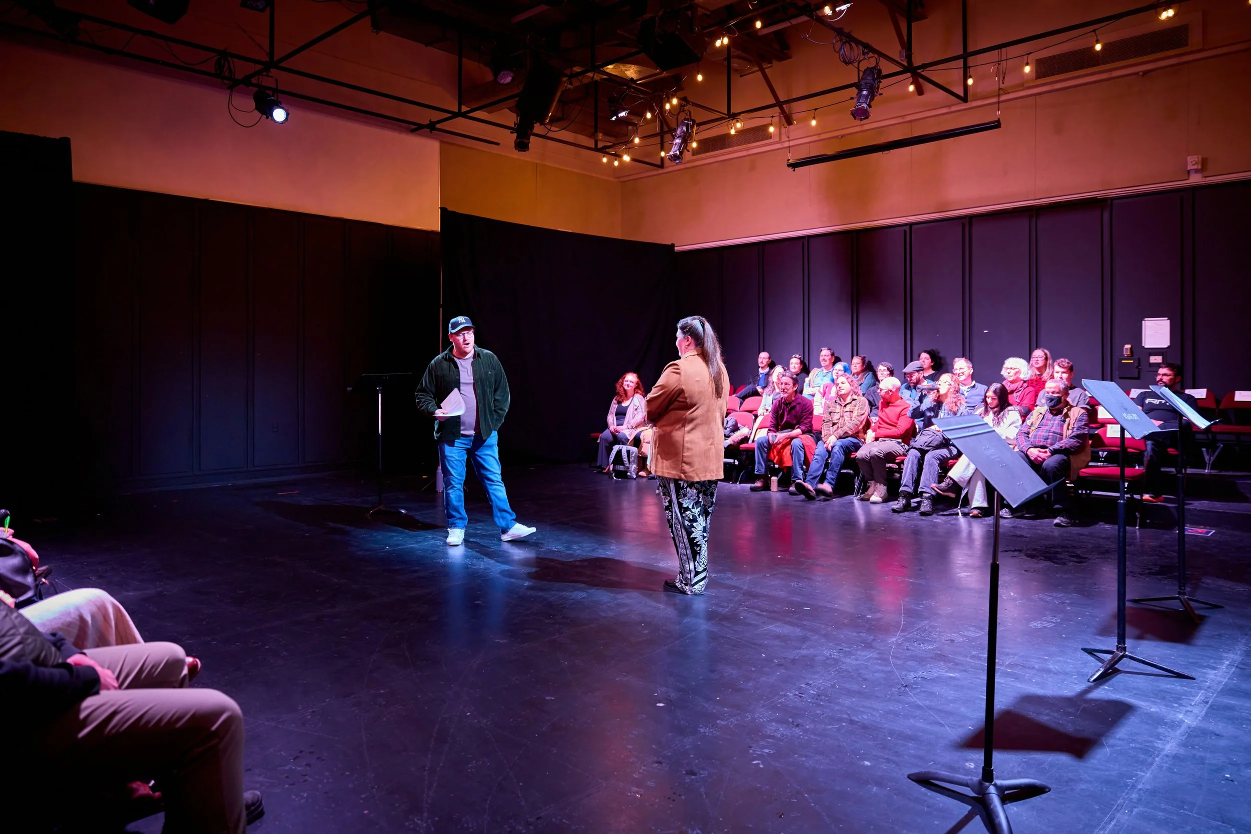 Une scène de théâtre ou de répétition avec deux personnes au centre, une parlant et l'autre écoutant, entourées d'une audience assise dans un espace avec un plafond avec des lumières suspendues.
