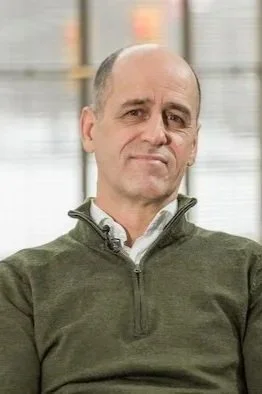 Homme d'âge moyen avec une expression neutre, portant un pull vert foncé et une chemise blanche, en intérieur.