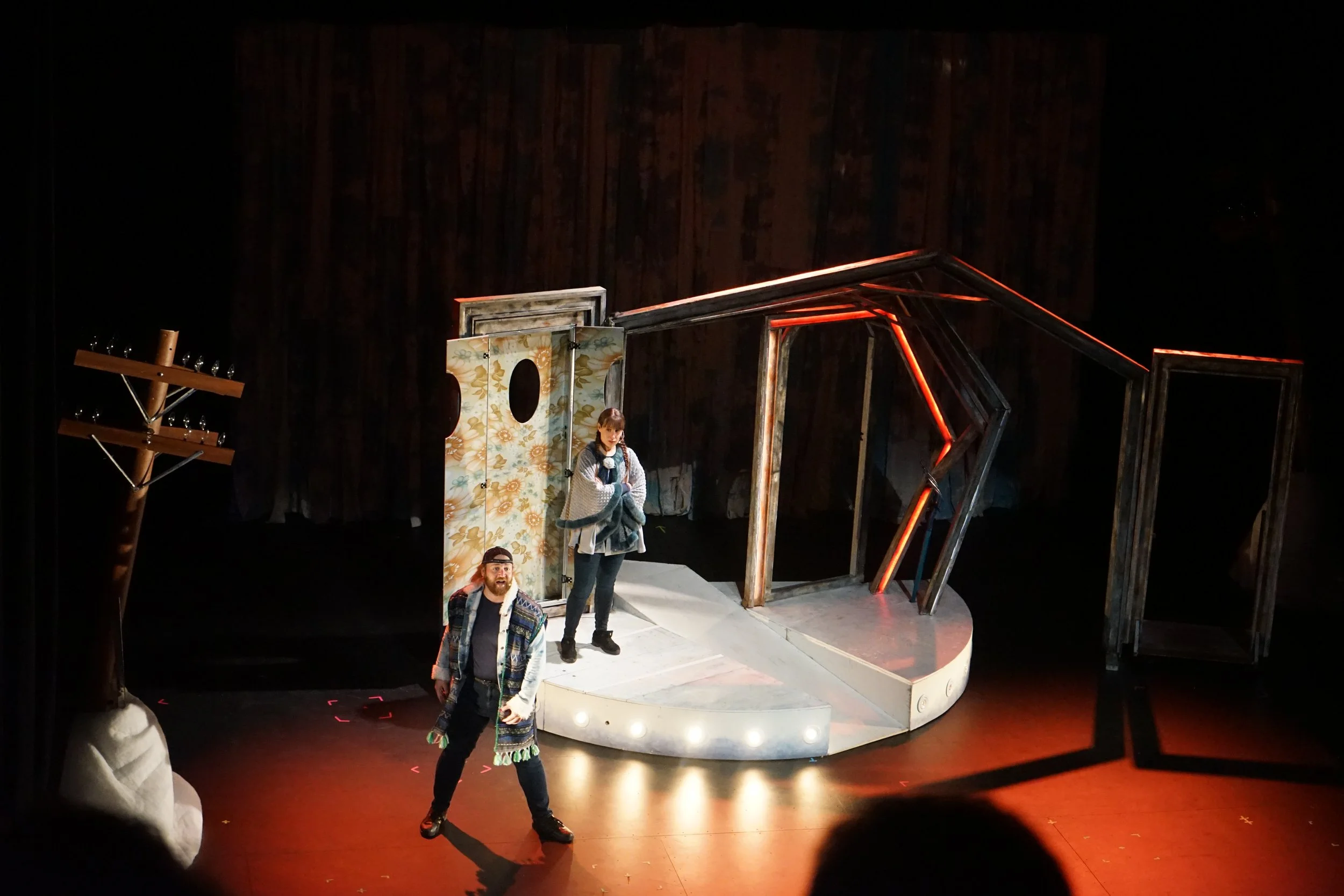 Scène de théâtre avec deux acteurs sur une scène comprenant une structure en bois, un panneau décoratif et un arbre en bois, dans un décor sombre.