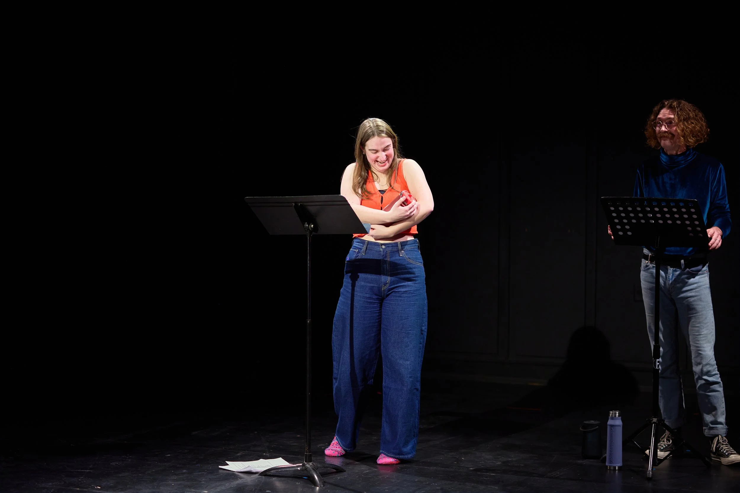 Deux personnes sur scène, une femme en débardeur orange et pantalon en jean, riant et tenant un objet rouge contre sa poitrine. À côté, un homme avec des cheveux bouclés, portant un pull bleu velours et un pantalon gris, souriant et debout derrière u