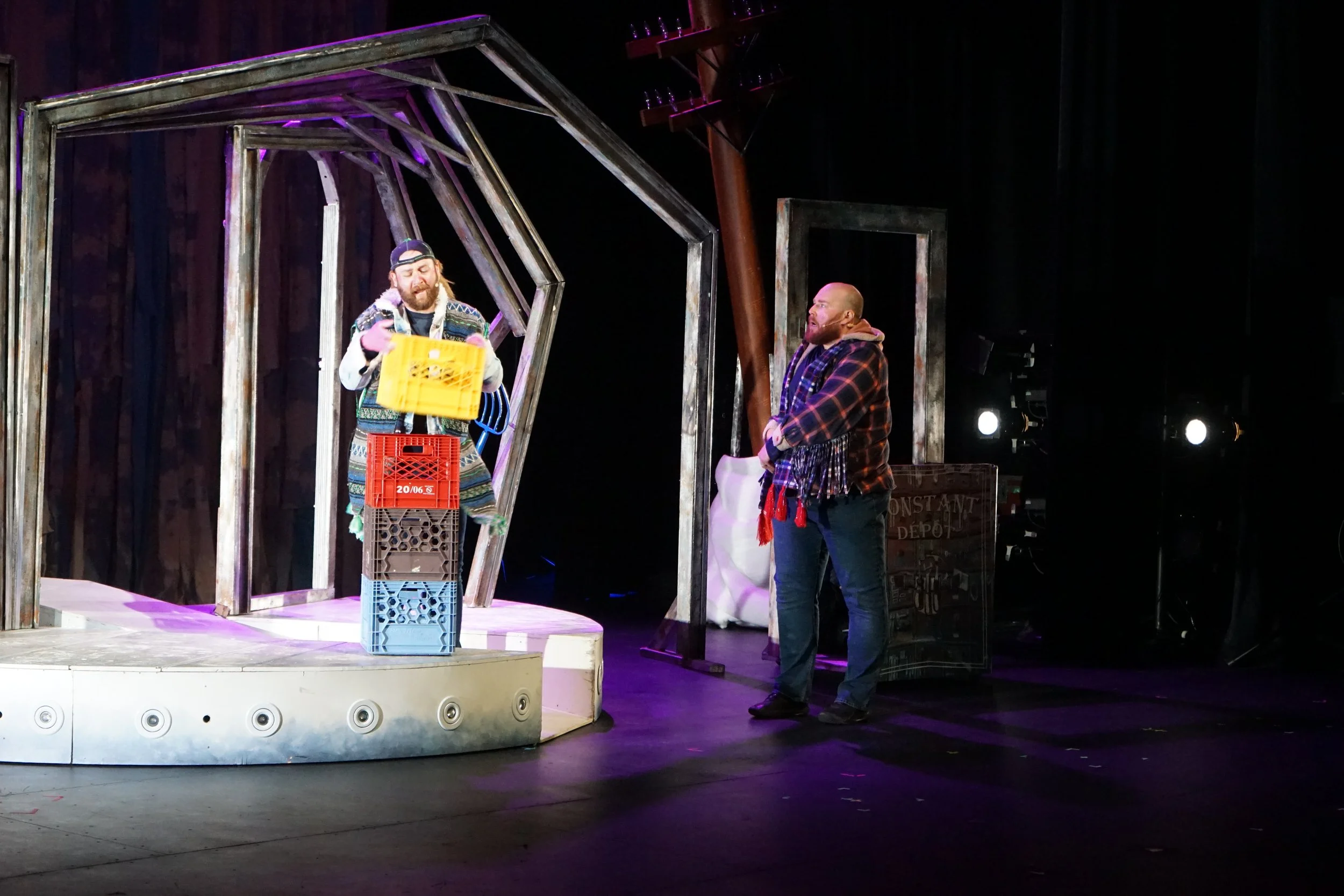 Deux hommes sur scène, un sur un petit podium en arrière-plan, l'autre au sol. L'homme sur le podium tient une caisse en plastique jaune, sourit, et porte des vêtements colorés et un chapeau. L'autre homme porte un manteau à carreaux et regarde l'aut
