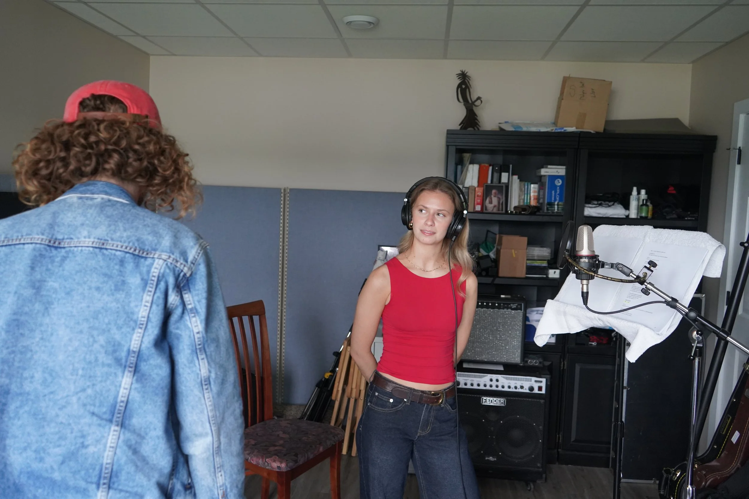 Une jeune femme portant un haut rouge et un jean discute avec un homme portant une veste en jean et une casquette rouge, dans une salle avec équipement d'enregistrement et un canapé. 