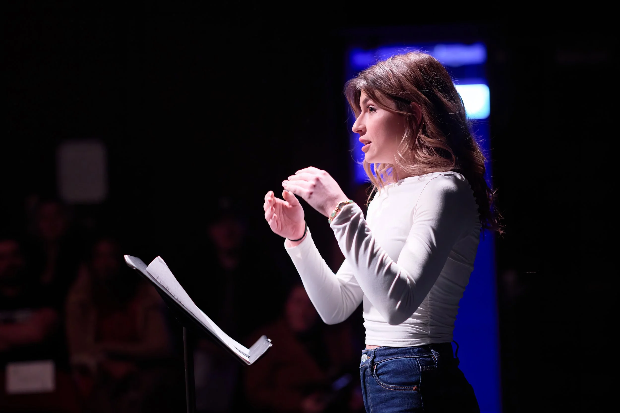 Une femme blonde parle lors d'une présentation ou d'un discours, avec une audience en arrière-plan dans une pièce sombre, devant un pupitre avec des notes.