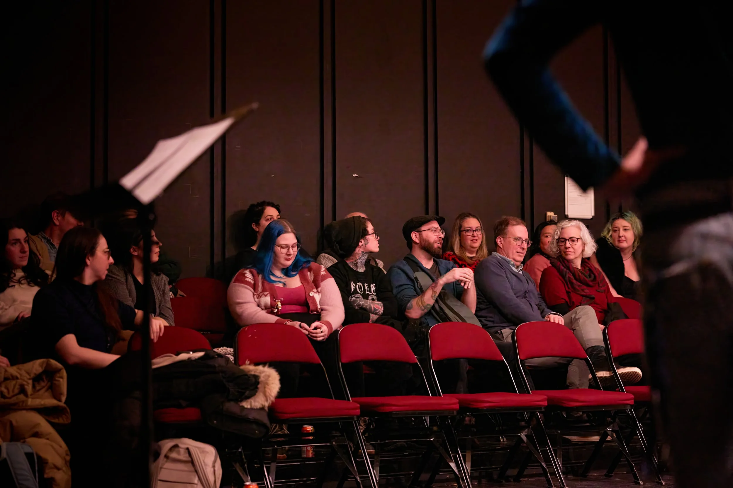 Auditoire de personnes assises dans une salle, certains regardant vers l'avant, d'autres discutant, avec des chaises rouges.