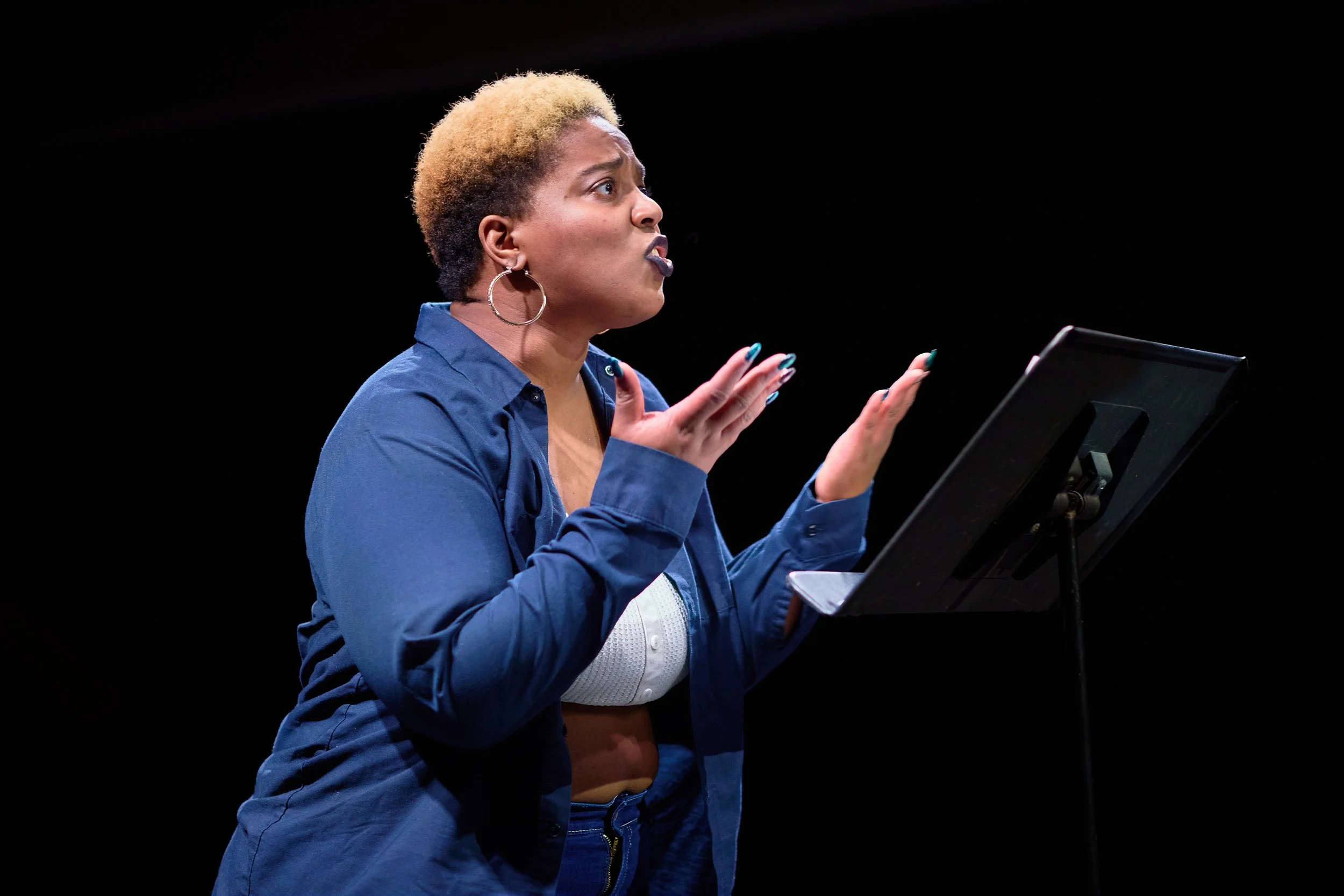 Femme parlant devant un pupitre, faisant un geste expressif, vêtue d'une chemise bleue, avec des cheveux courts et un fond noir.