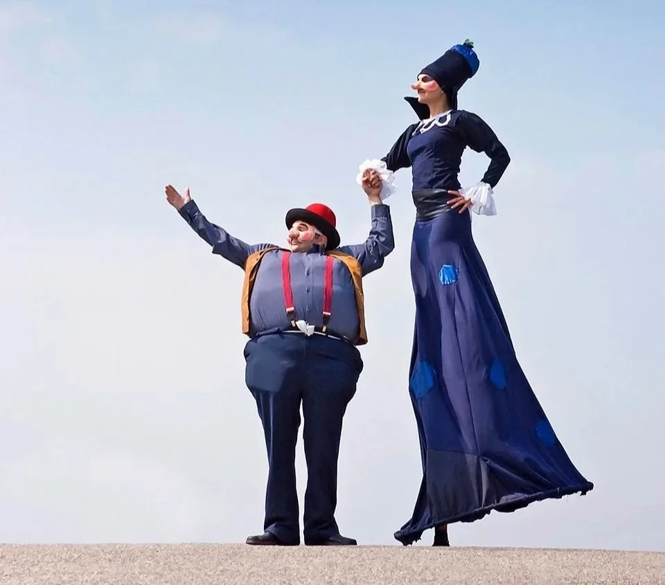 Deux mimes en costumes colorés en train de jouer, l'un avec une grosse personne et l'autre avec une longue personne sur un fond de ciel bleu.