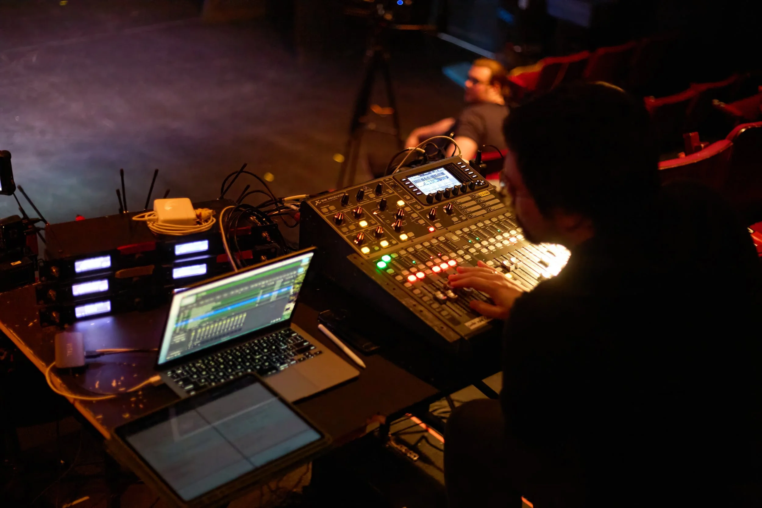Un DJ utilisant une console de mixage dans une salle de concert sombre avec des spectateurs assis en arrière-plan.