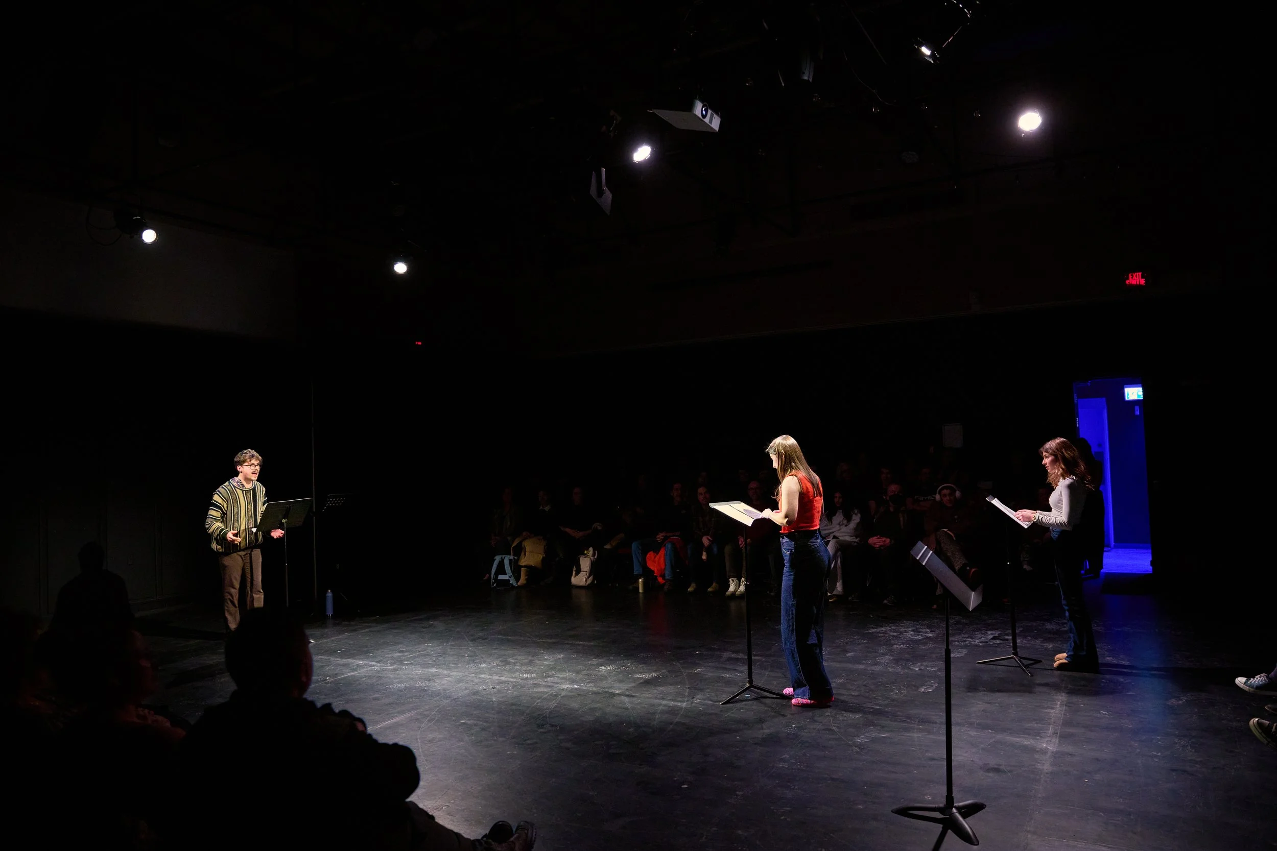 Trois personnes sur une scène de théâtre, deux femmes et un homme, en train de lire ou réciter, avec un public assis dans l'ombre. La scène est sombre, avec un éclairage ciblé sur les intervenants.