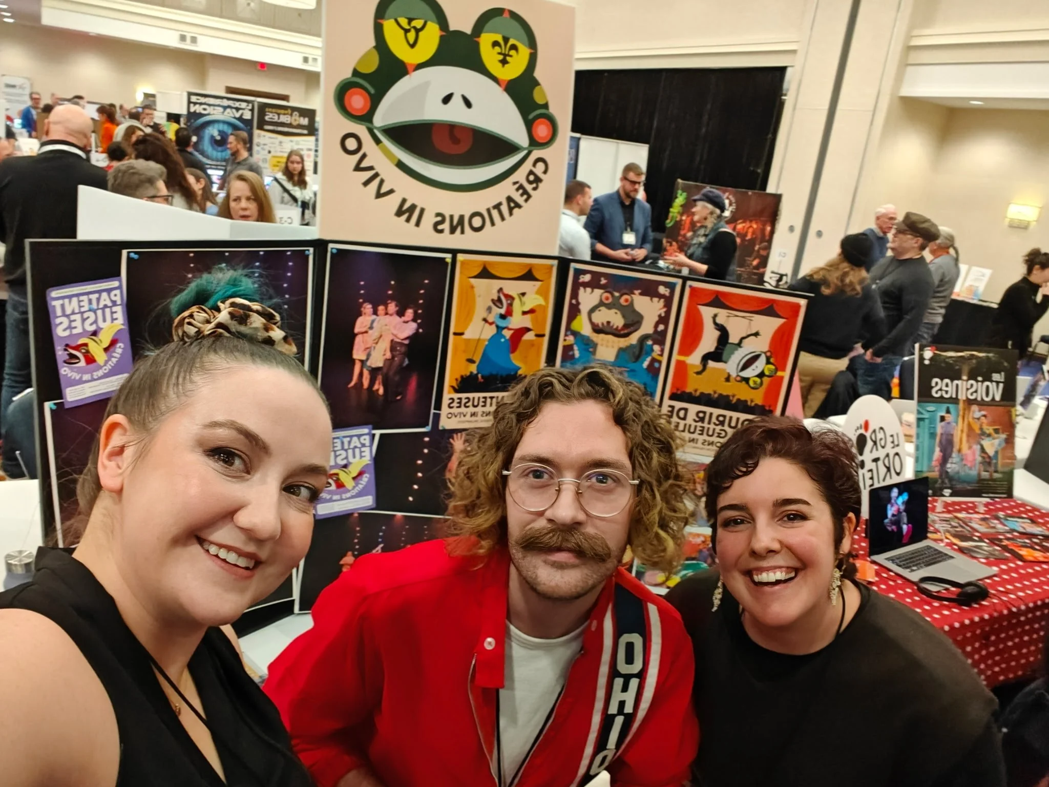 Créations In Vivo. Trois personnes posent pour une photo lors d'une convention. Derrière eux, il y a des affiches colorées avec des illustrations de spectacles ou de personnages animés, et d'autres visiteurs dans un grand hall.