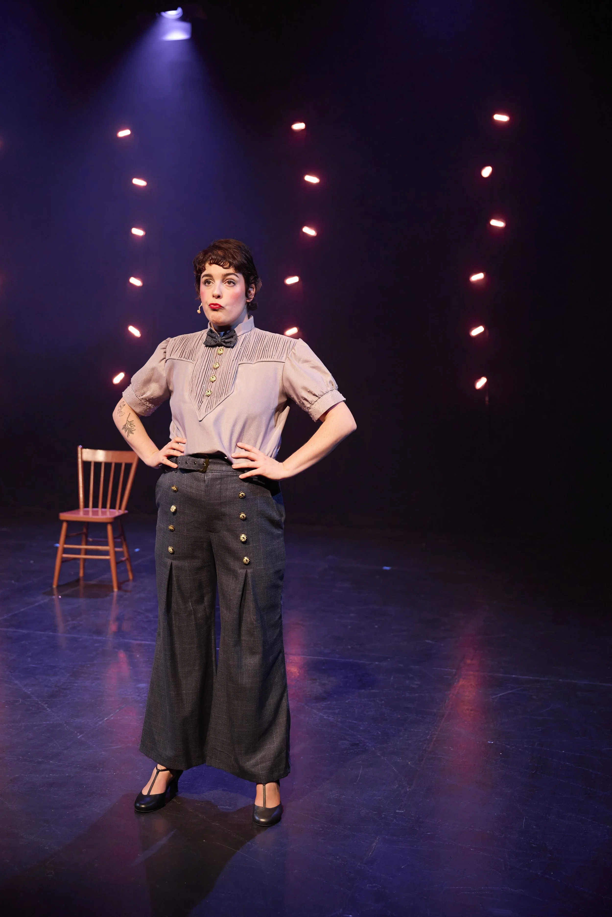 Une femme avec des vêtements vintage sur scène, avec une chaise en bois derrière elle et des lumières colorées en arrière-plan.