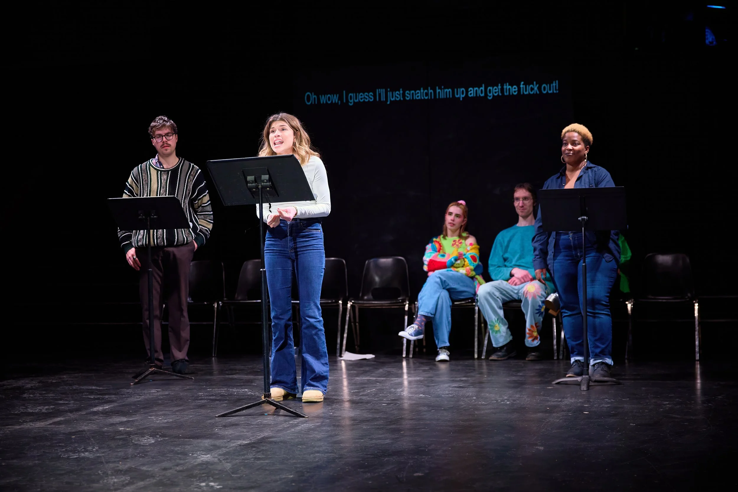 Groupe de personnes en scène, la femme au centre parle. Il y a une projection avec du texte en anglais derrière eux.