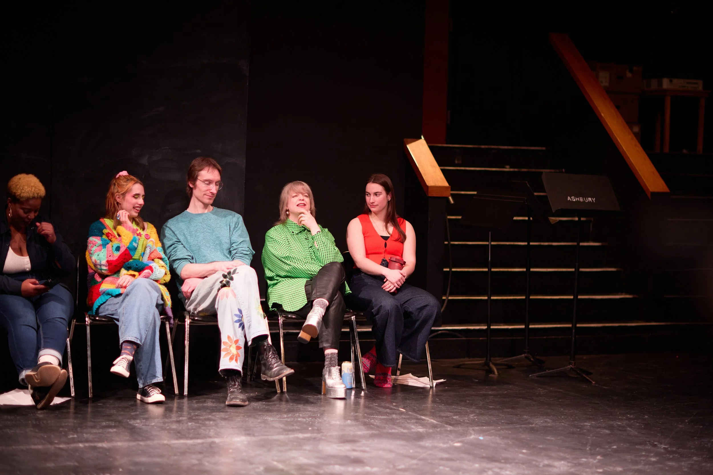 Cinq personnes assises sur une scène, discutant ou participant à une activité. La scène est sombre avec un piano en arrière-plan et des stands de musique.
