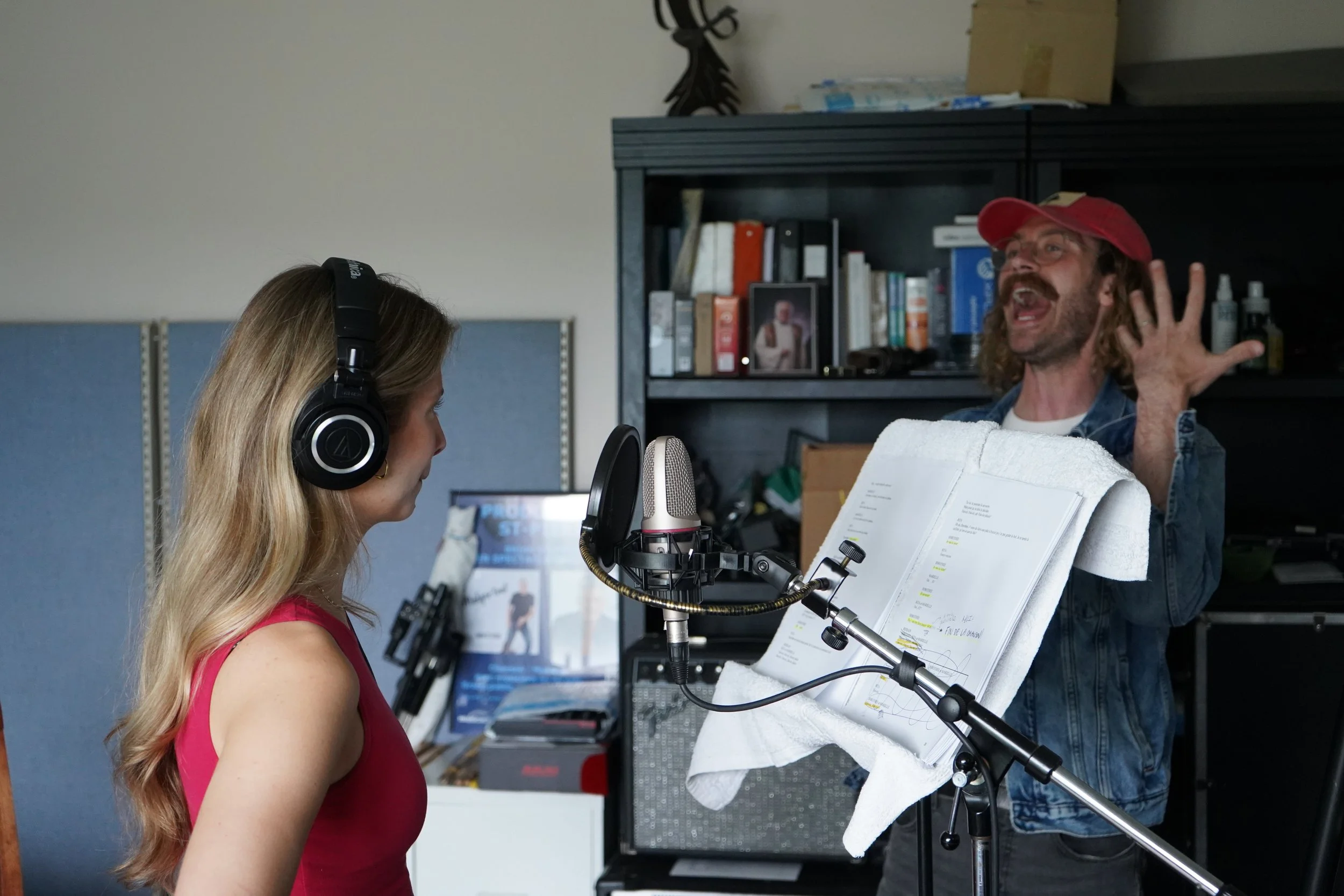 Une femme en studio d'enregistrement, portant un casque audio et une robe rouge, chante ou parle dans un microphone. Un homme avec une barbe, des cheveux longs et portant une casquette rouge, semble répondre ou discuter avec elle, tout en levant la m