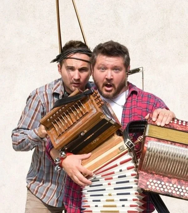 Deux hommes portant une panoplie d'instruments de musique, dont un accordéon, un piano à quenouilles, un clavier et un autre instrument, avec des expressions de surprise ou d'étonnement, posant contre un mur blanc.