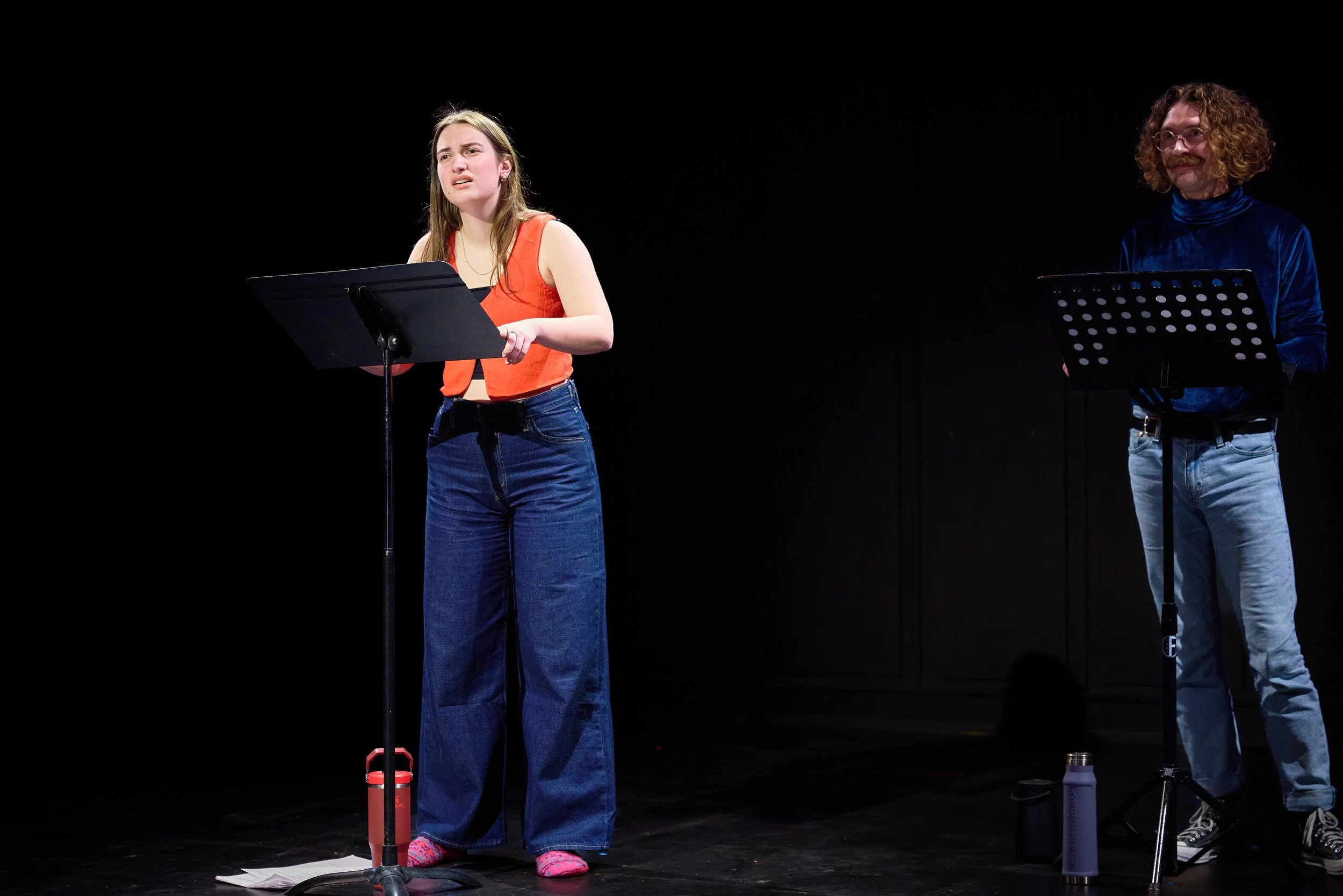 Deux personnes participent à une lecture ou une performance sur scène, chacune derrière un pupitre, dans un espace sombre.