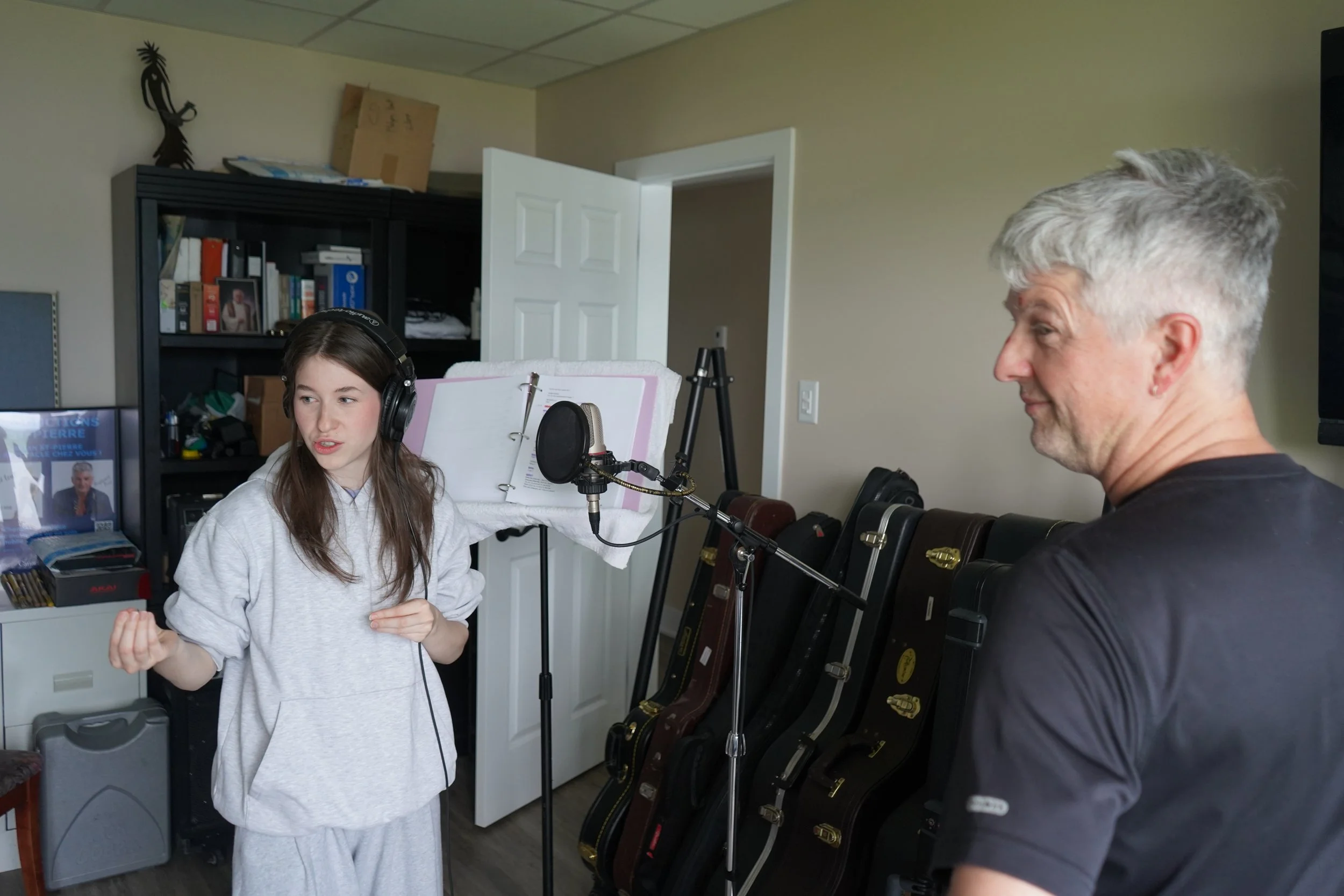 Une jeune fille avec un casque, parlant dans un microphone, une femme plus âgée à côté, dans une pièce avec étagère contenant des livres et des instruments de musique.