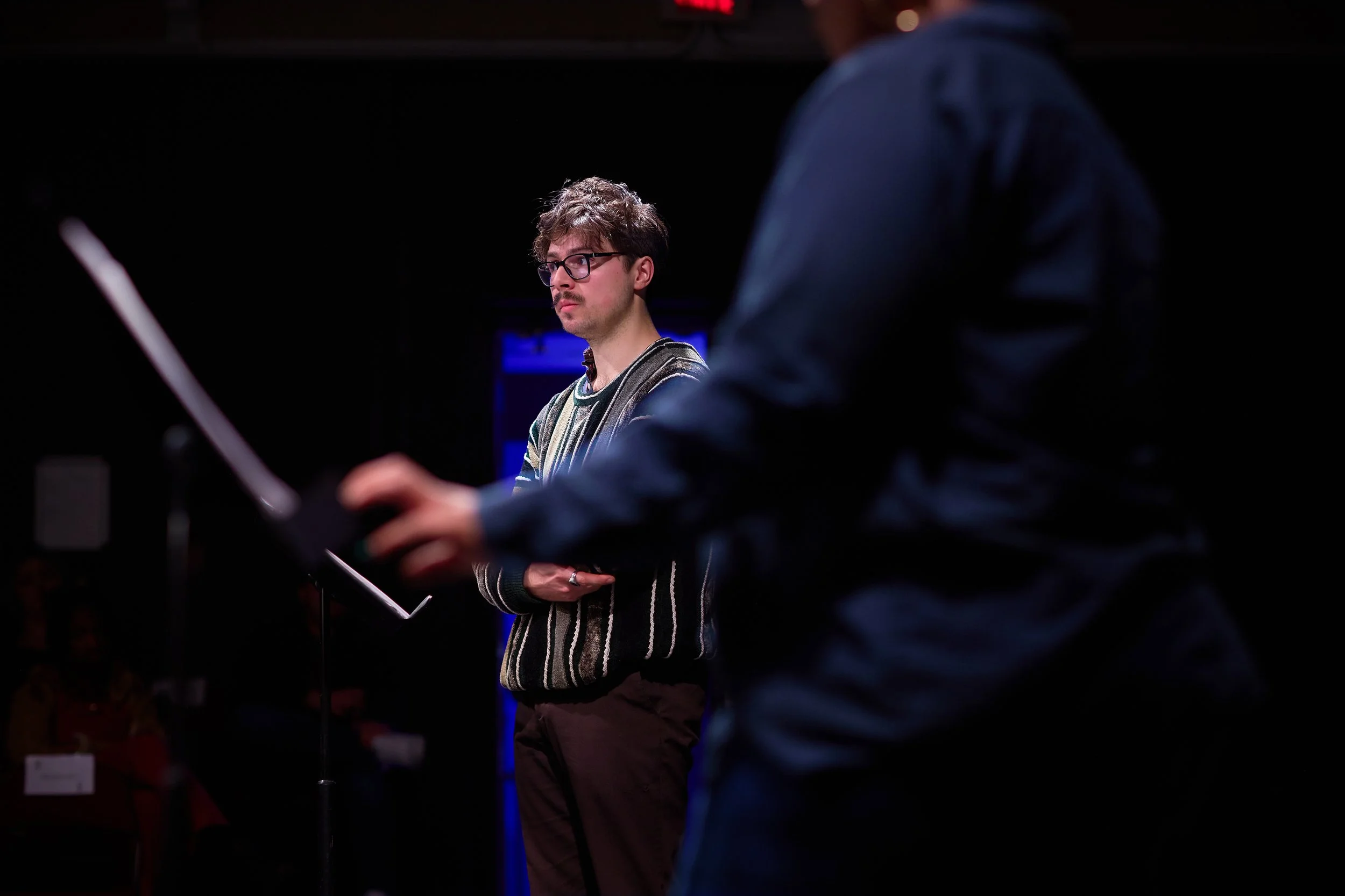 Un homme avec des lunettes et un pull rayé, debout devant un pupitre, dans une pièce sombre, probablement en train de lire ou de répéter pour une performance.
