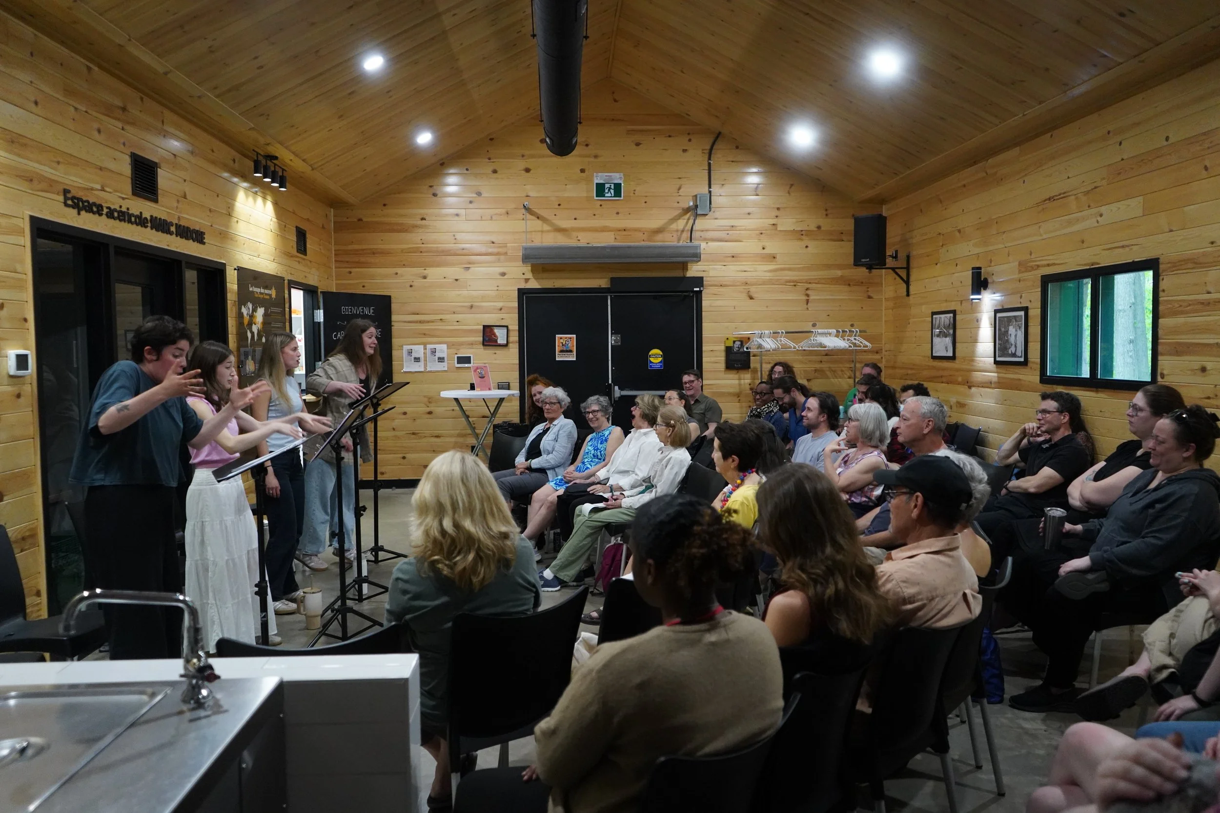 Une salle avec des personnes assises regardant une performance ou présentation. À gauche, un groupe de jeunes femmes chanteuses ou oratrices, avec un microphone. Les murs et le plafond en bois donnent une ambiance chaleureuse. Il y a un public varié,