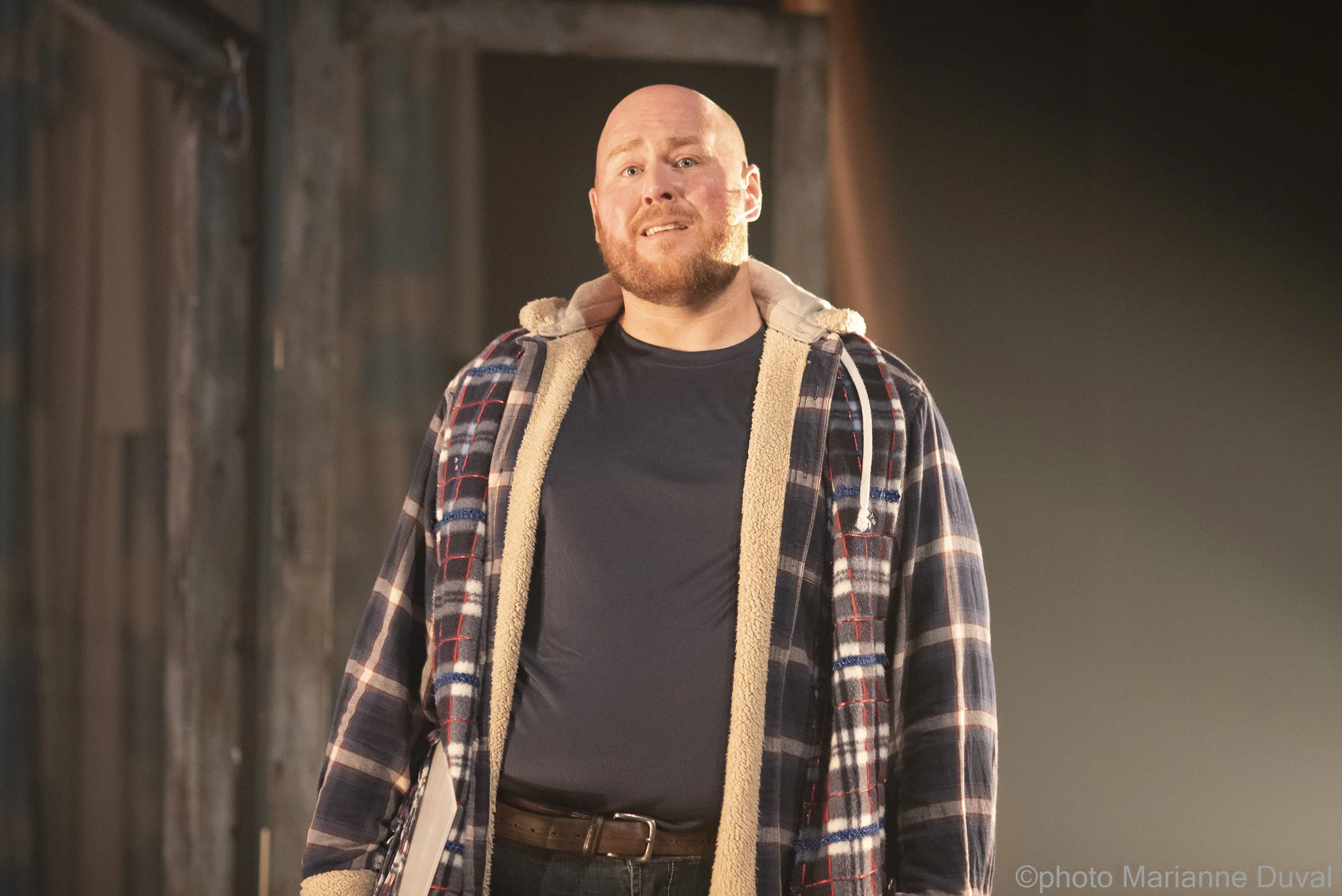 Un homme avec une barbe portant un sweat à capuche, un t-shirt noir, une veste à carreaux et tenant un cahier ou un livre, semble exprimer de l'inquiétude ou de la confusion.