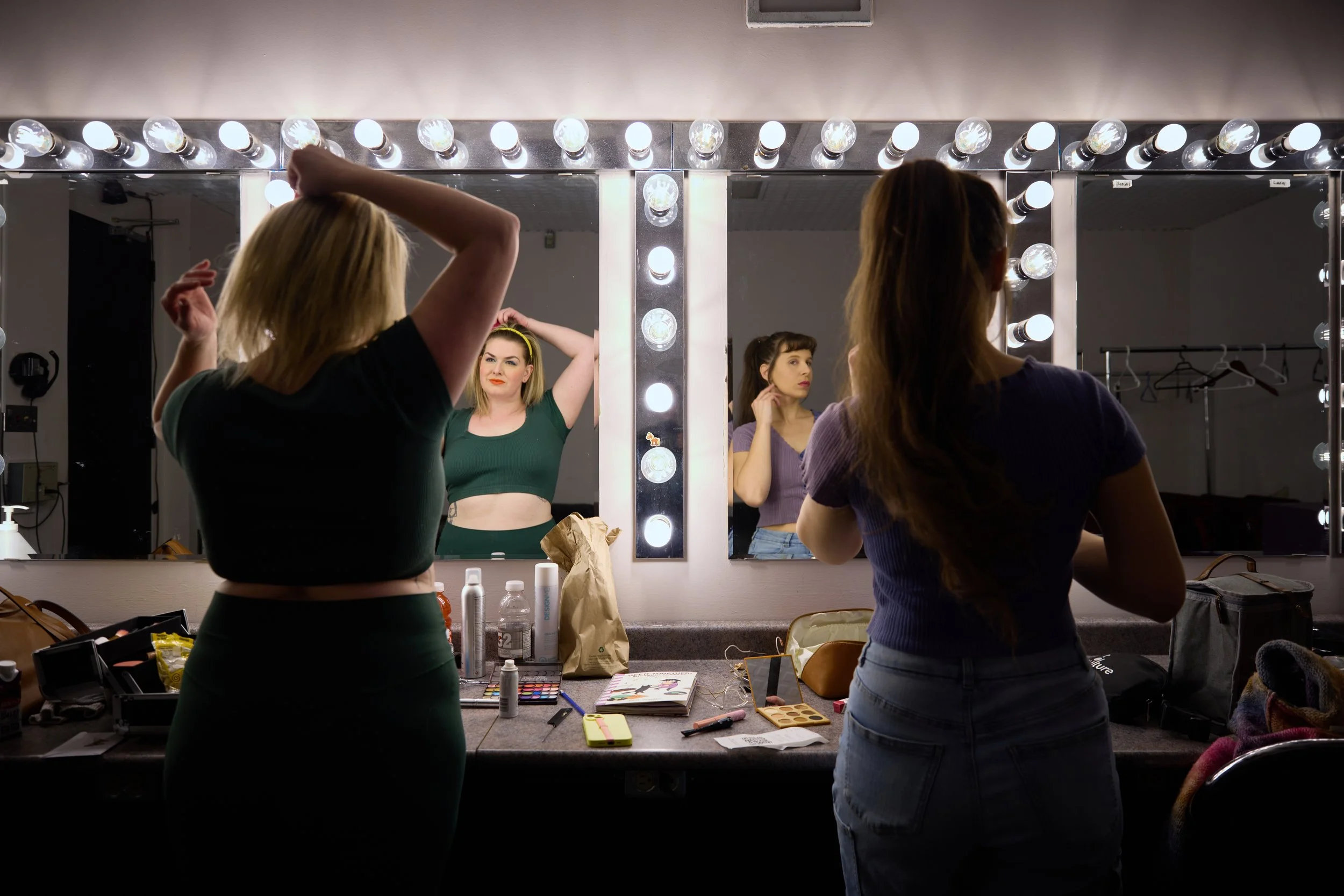 Deux femmes se regardent dans un miroir entouré d'ampoules, une préparant son maquillage et l'autre ajustant ses cheveux, dans une loge de maquillage.