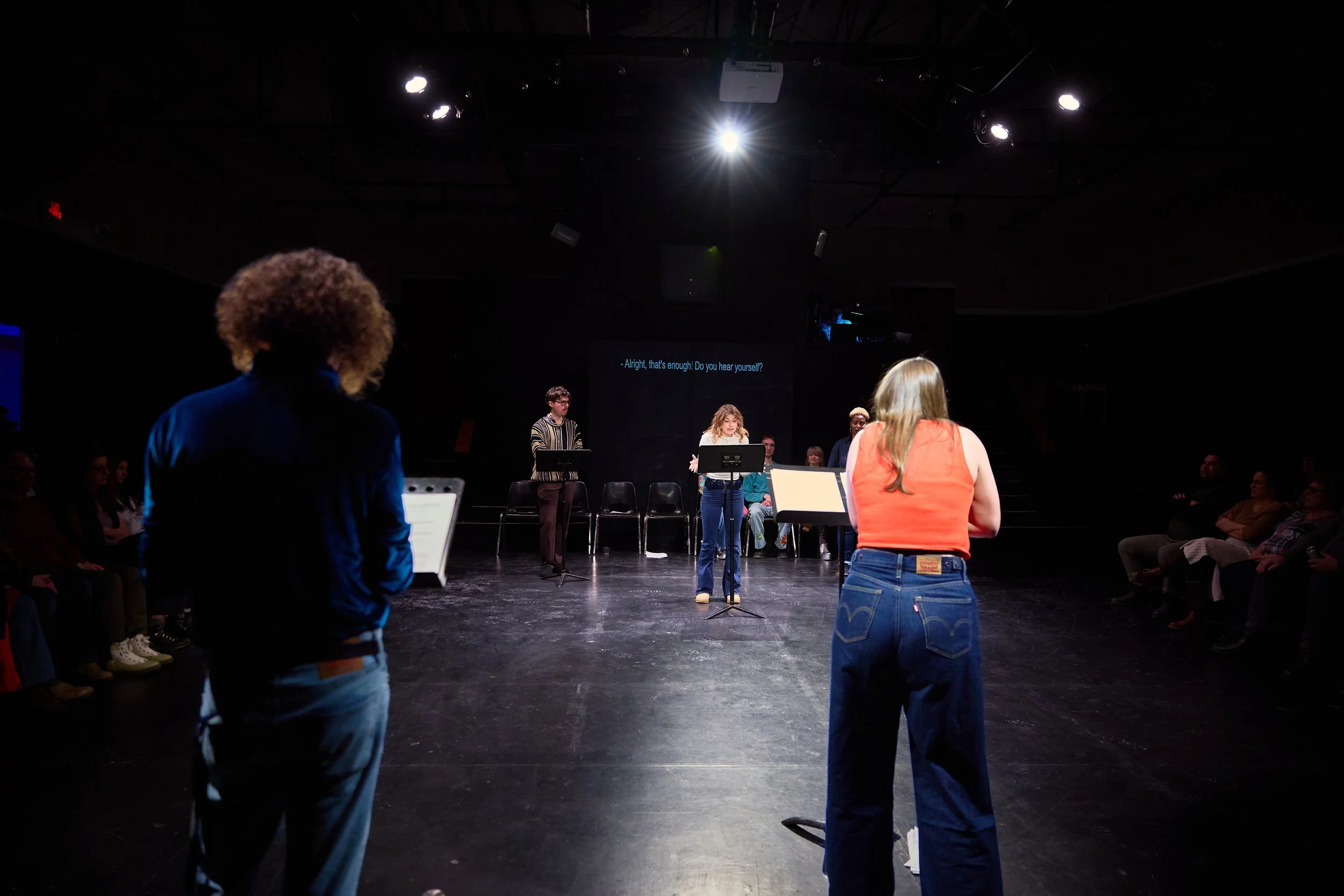 Scène de théâtre avec quatre acteurs en répétition sur scène devant un public, sous un éclairage intense.