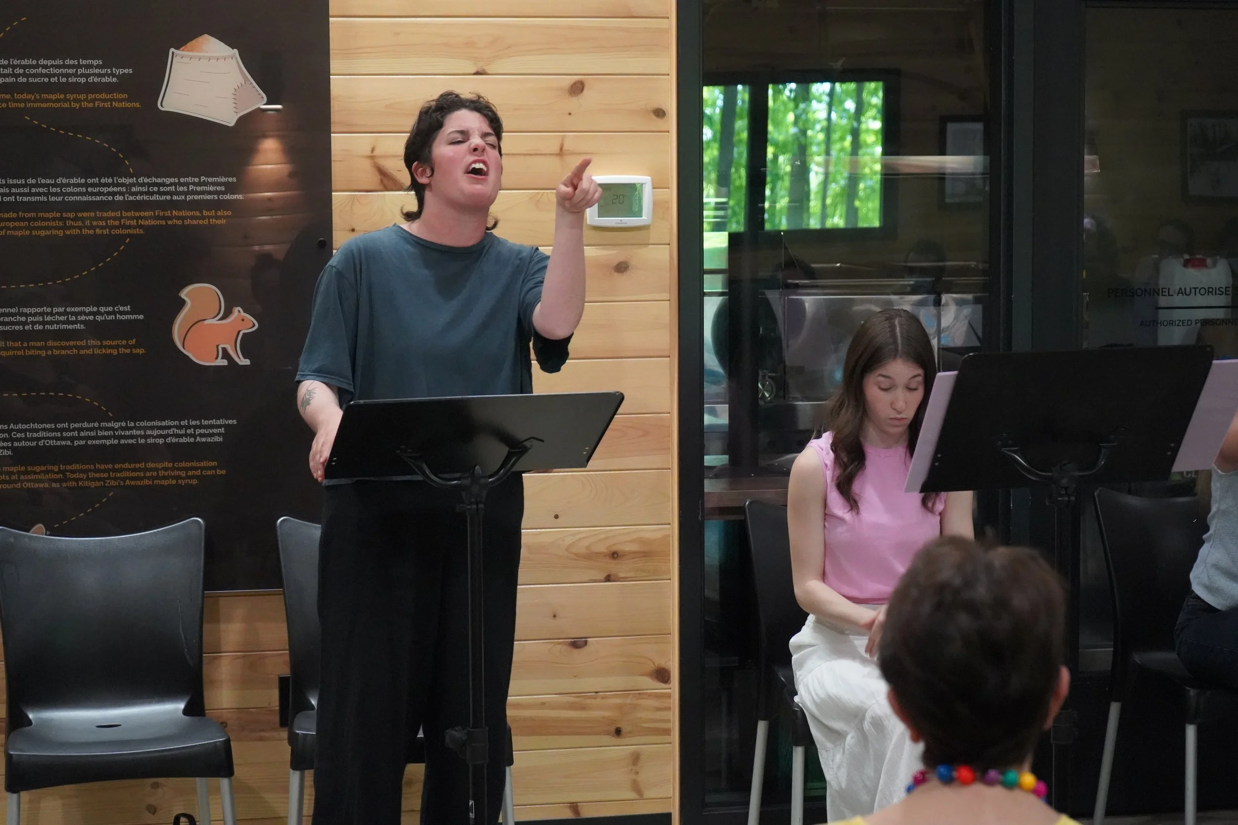 Une femme parle ou chante devant un micro dans un environnement intérieur en bois, avec d'autres personnes assises ou debout, dans une ambiance de groupe ou de classe. Une affiche pédagogique est visible derrière elle.