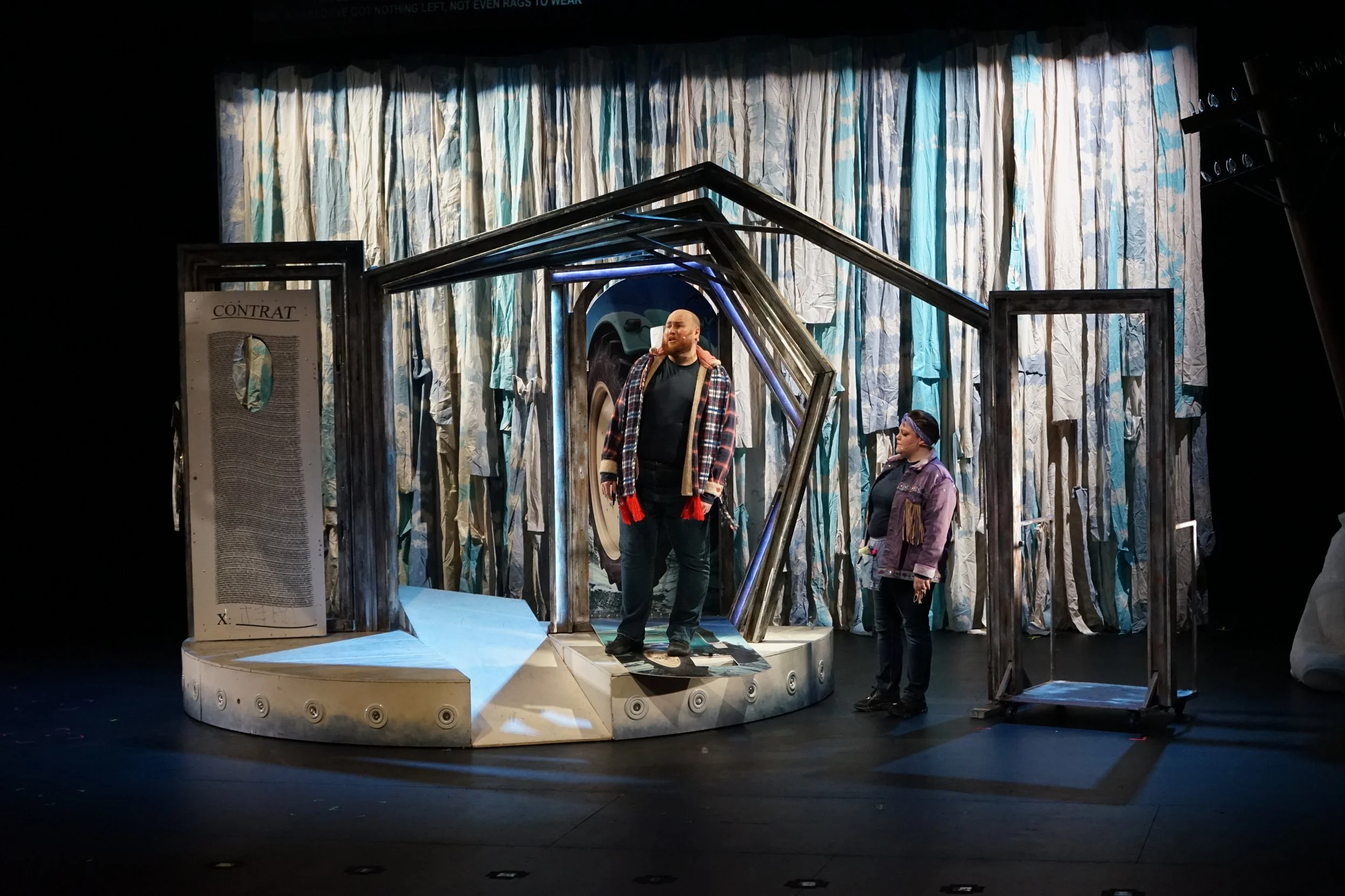 Scène de théâtre avec deux acteurs sur un plateau décoré d'une porte, d'un panneau 'Contrat', et d'un arrière-plan en tissu de couleurs bleues et blanches, une femme debout regarde un homme qui se tient sur une planche de skateboard.