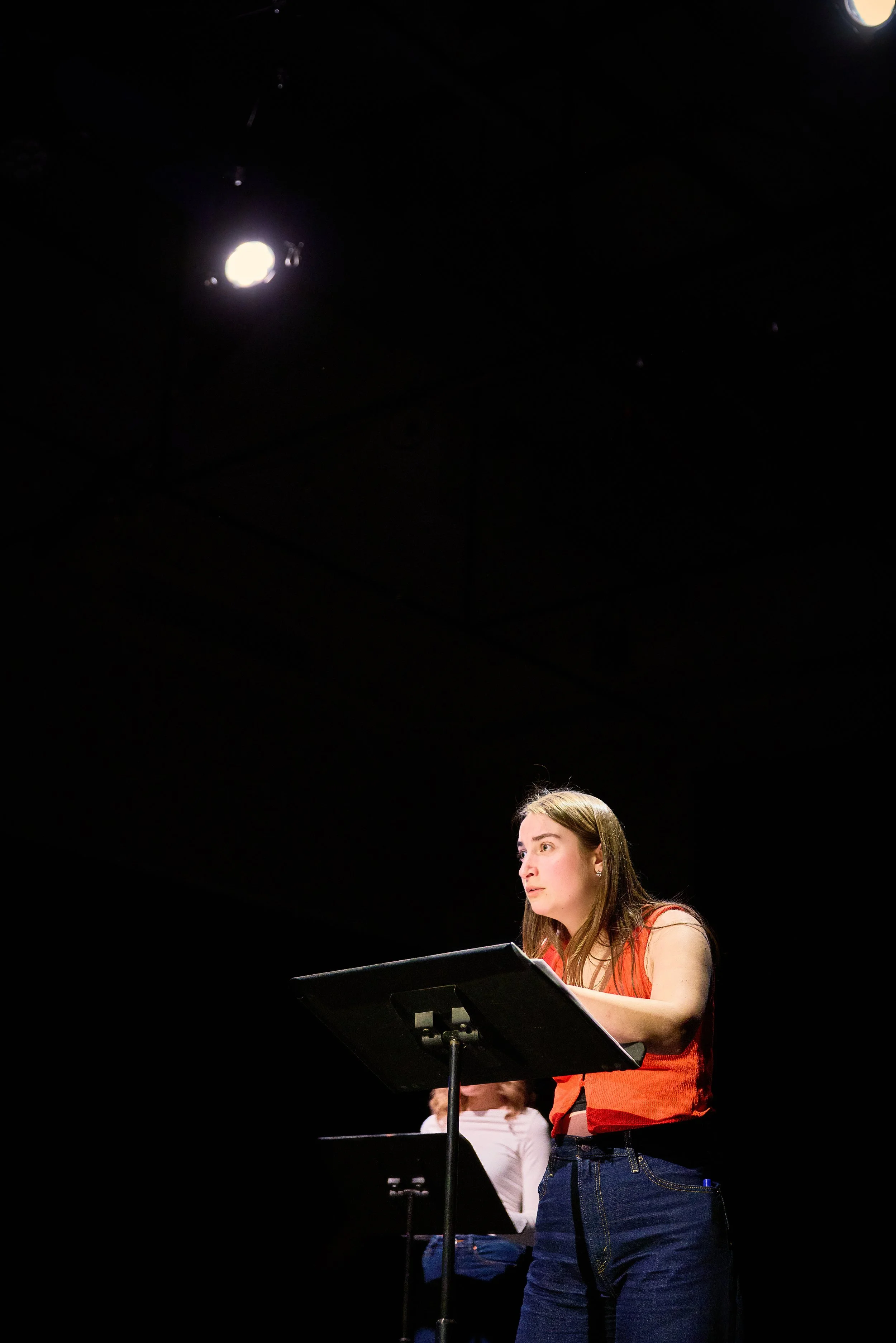 Une jeune femme avec des cheveux longs et une chemise sans manches rouge, lisant d'une partition sur un pupitre lors d'une répétition ou d'un concert. Il y a une lumière forte au plafond dans un espace sombre.