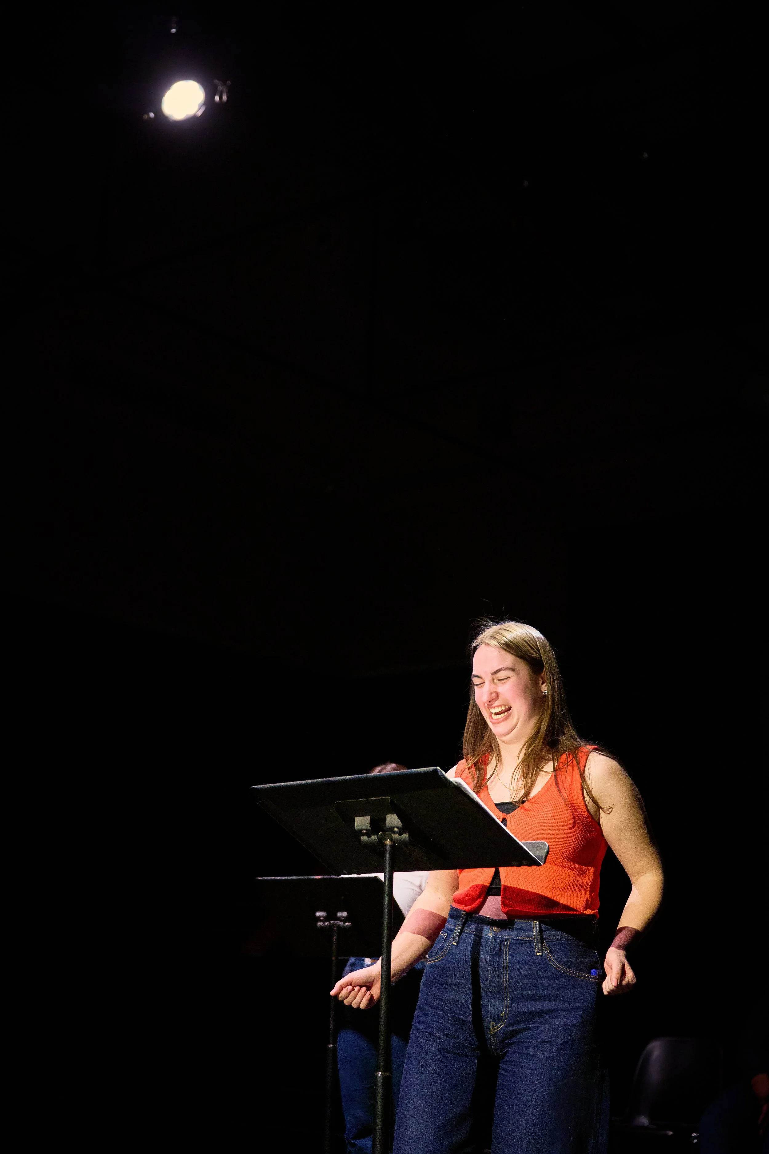 Une jeune femme souriante, portant un haut rouge et un jean, se tient devant un pupitre avec une partition, sur scène. L'éclairage est concentré sur elle, le fond est sombre.