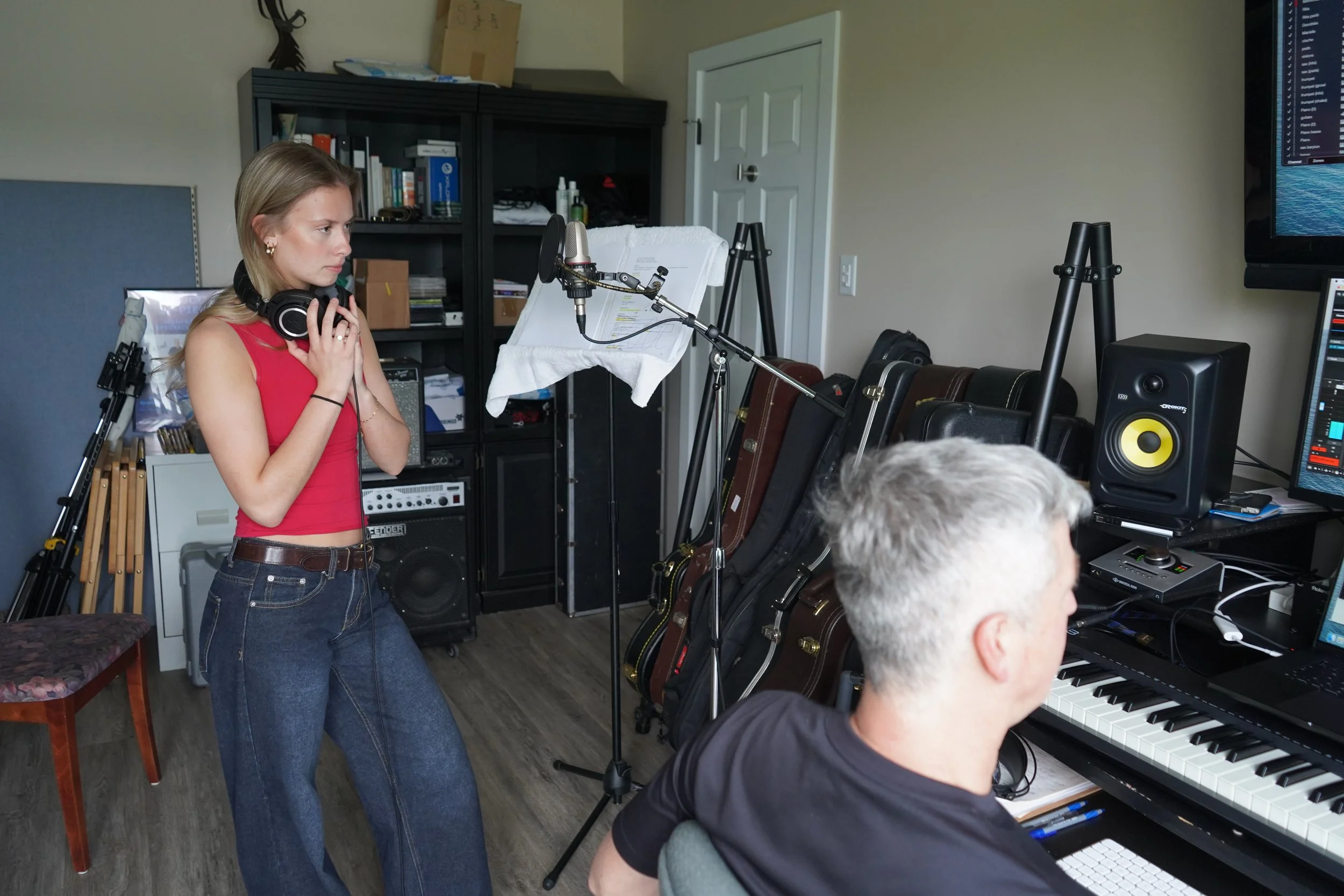 Une jeune femme avec des écouteurs près du cou se tient à côté d'un homme jouant du piano dans une pièce avec de nombreux instruments de musique et du matériel d'enregistrement.
