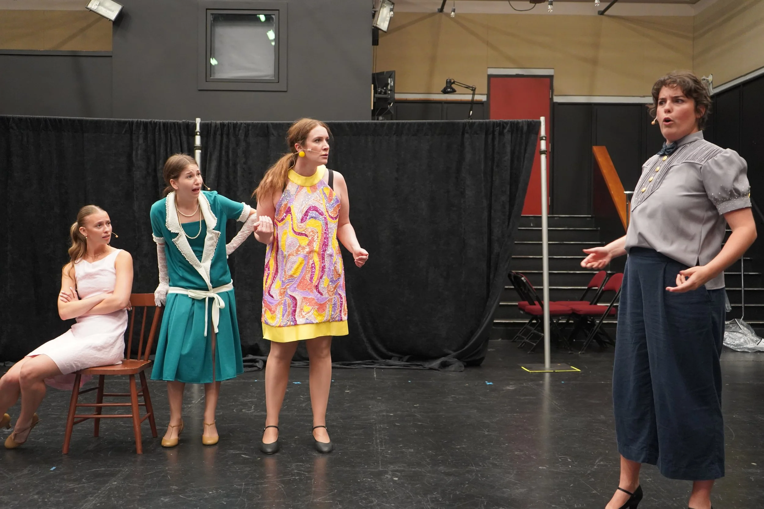 Quatre jeunes actrices en costume sur scène, en pleine performance, avec une scène noire en fond et des éclairages au plafond.