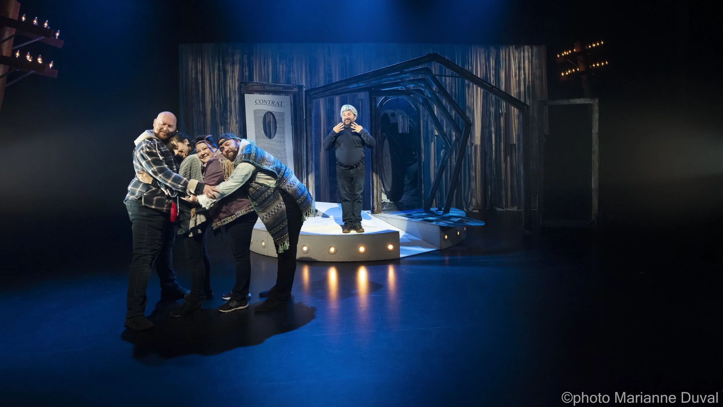 Groupe de cinq personnes en costume sur scène, l'une d'elles au centre avec un micro, autres qui se tiennent par la main, en arrière-plan un décor de théâtre avec panneau 'Contrat' et structure en bois, éclairage artistique.