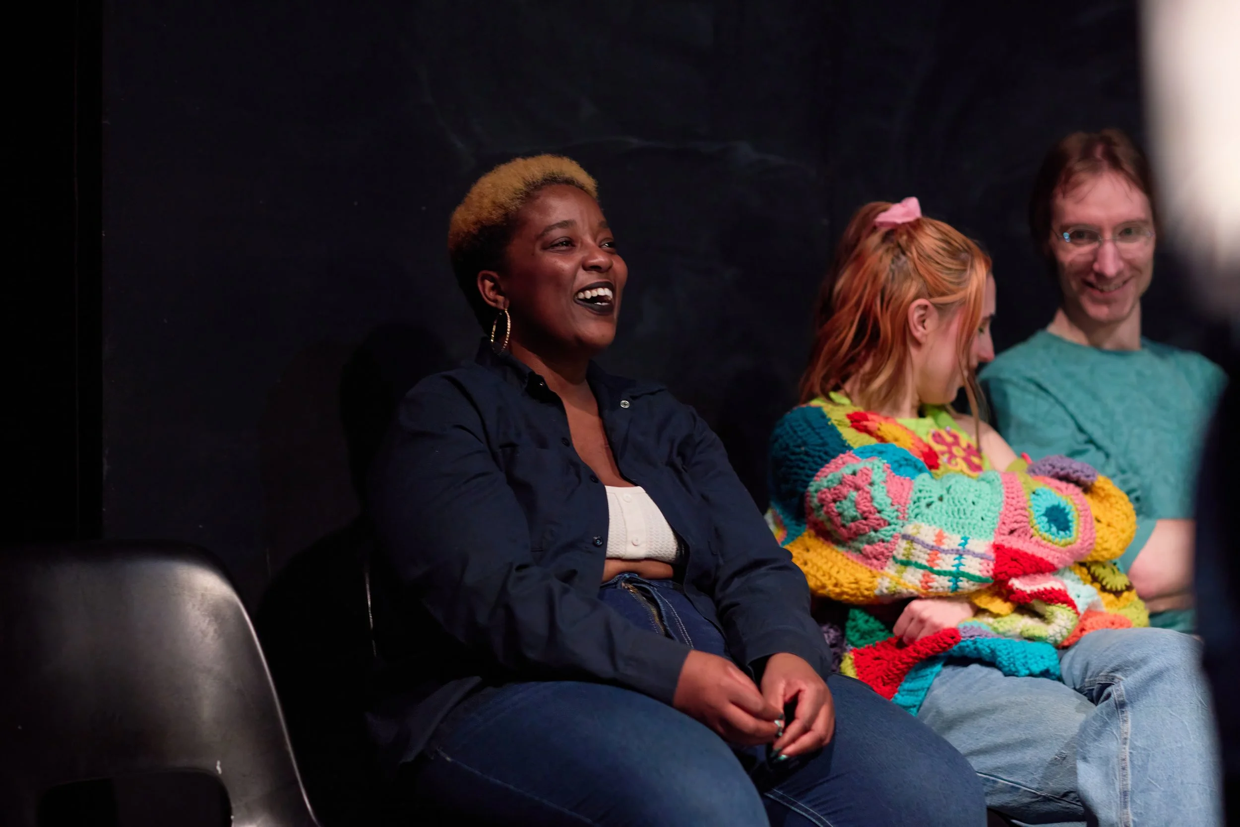 Trois personnes assises côte à côte, dont une femme noire qui sourit largement, et deux autres personnes, dont une fille avec un sweat coloré et un homme avec des lunettes, dans un environnement intérieur.