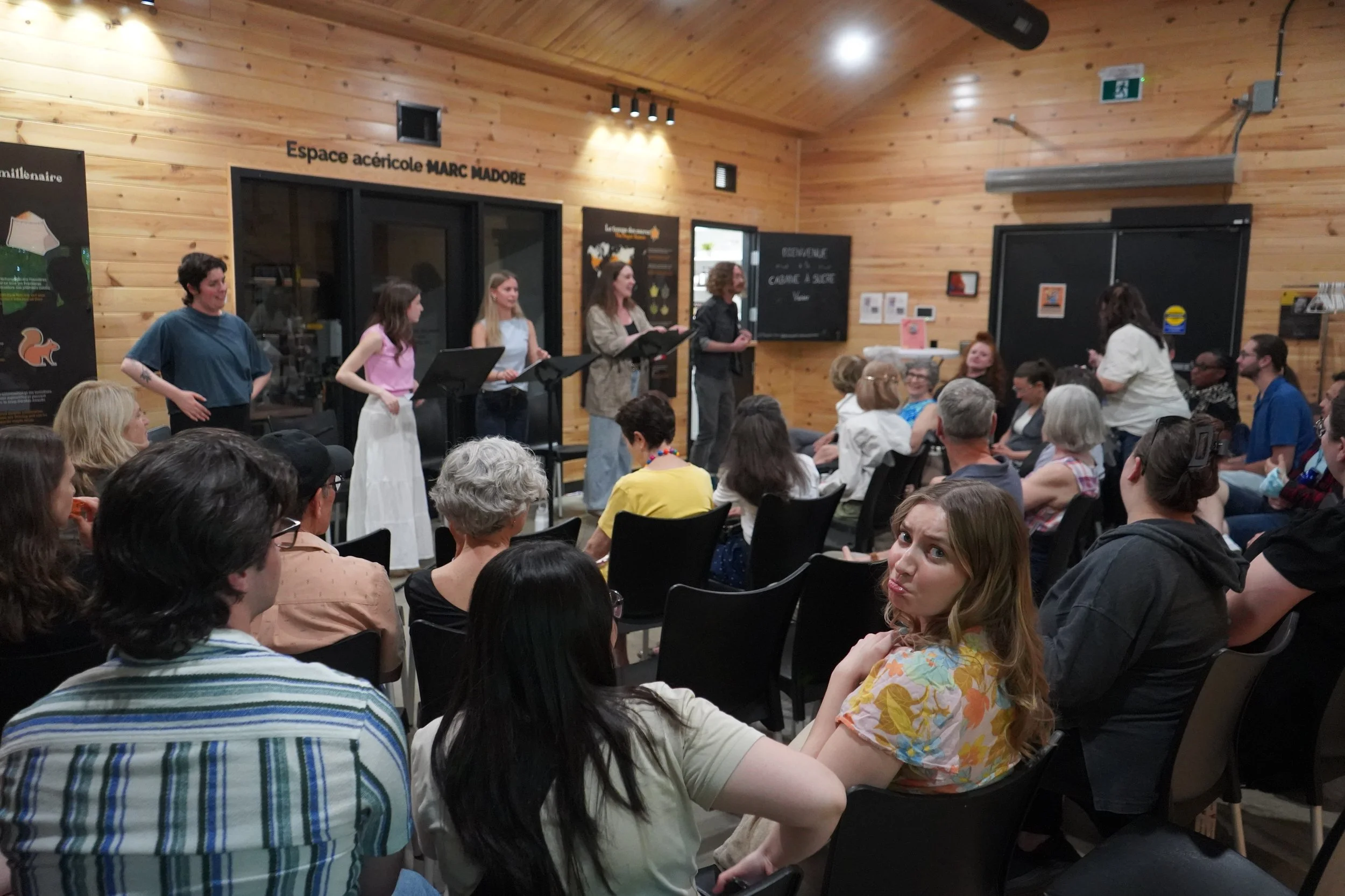 Une salle remplie de personnes assises et regardant un groupe de femmes qui chante ou parle sur une scène, dans un espace avec des murs en bois et des affiches sur le mur.