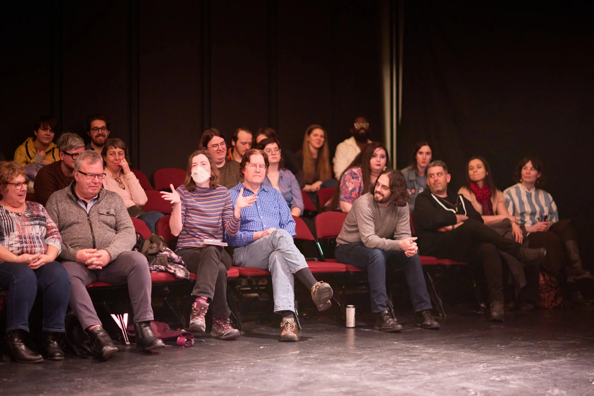 Groupe de personnes assises dans une salle de théâtre ou de conférence, attentives ou en train de participer à une discussion, avec certains souriants ou échangeant.
