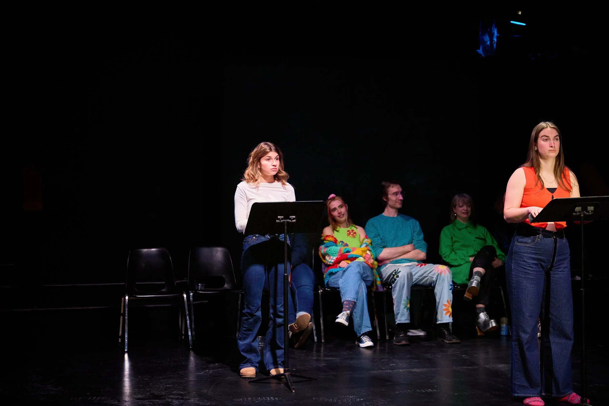 Jeune femme debout derrière un pupitre, parlant ou lisant, dans un espace scénique sombre, avec d'autres personnes assises derrière elle sur scène.
