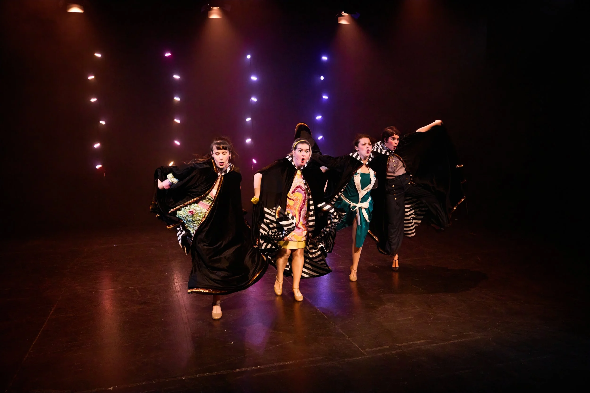 Quatre femmes en costume de théâtre sur scène, en train de faire une performance avec des expressions dramatiques et des costumes colorés, sous un éclairage artistique avec spots lumineux en arrière-plan.