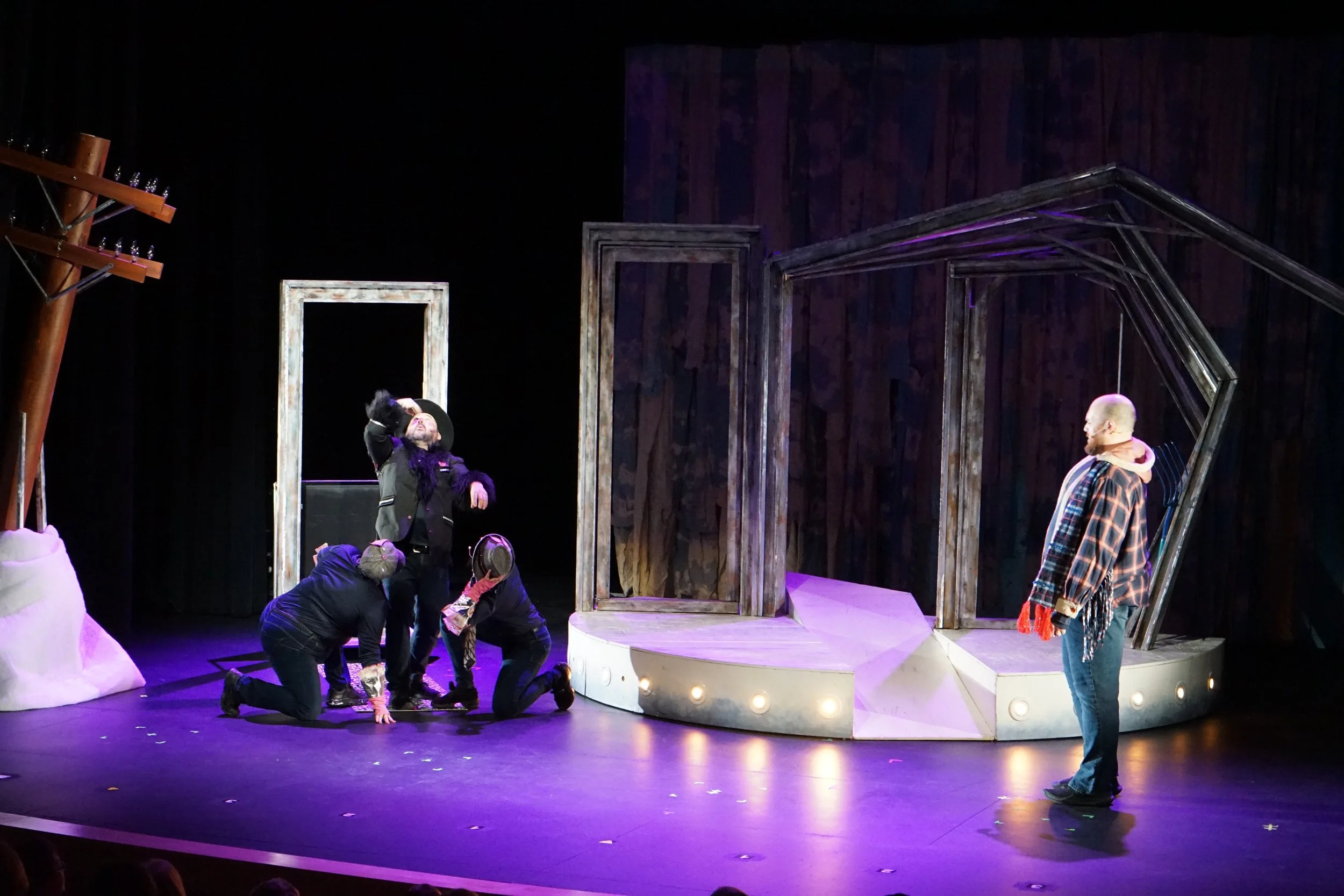 Acteurs de théâtre sur scène, certains à genoux, en train de jouer une scène dramatique, avec décor de fenêtre et structure en bois, sous éclairage violet.