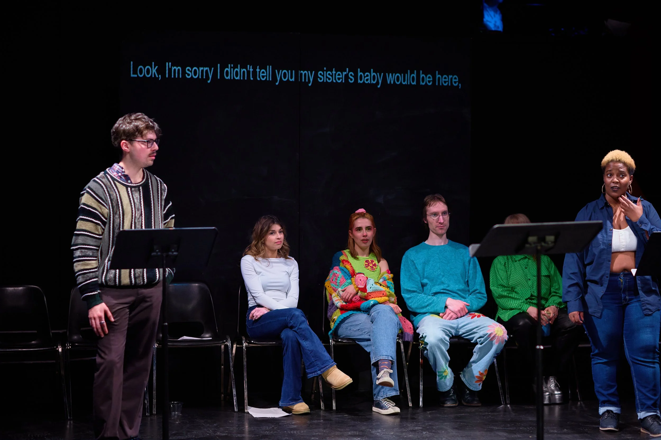 Groupe de personnes lors d'une lecture ou performance sur scène, avec un texte en anglais en arrière-plan et plusieurs personnes assises ou debout, une femme s'exprimant.
