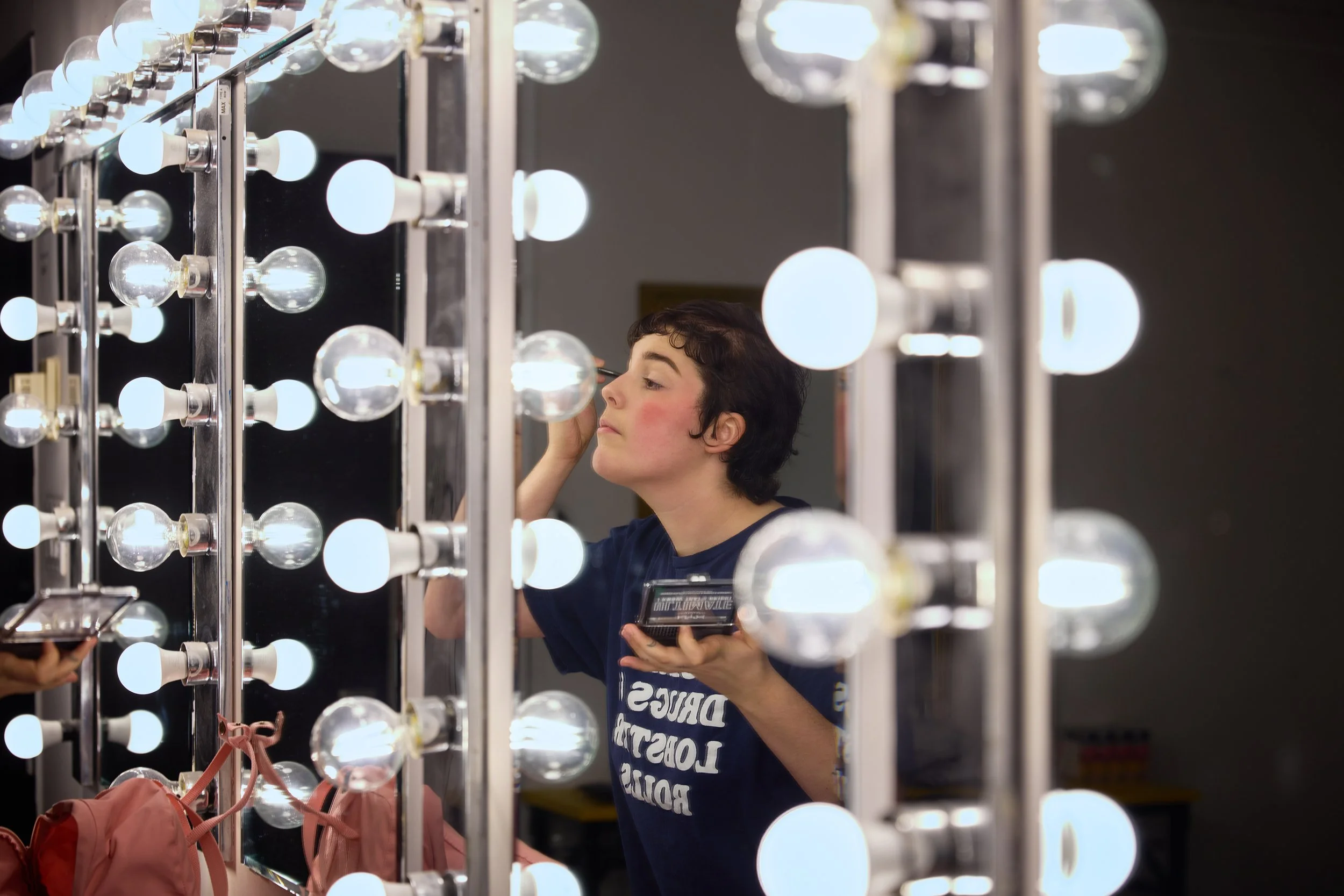 Une jeune personne maquillée, observant son reflet dans un miroir entouré d'ampoules lumineuses.